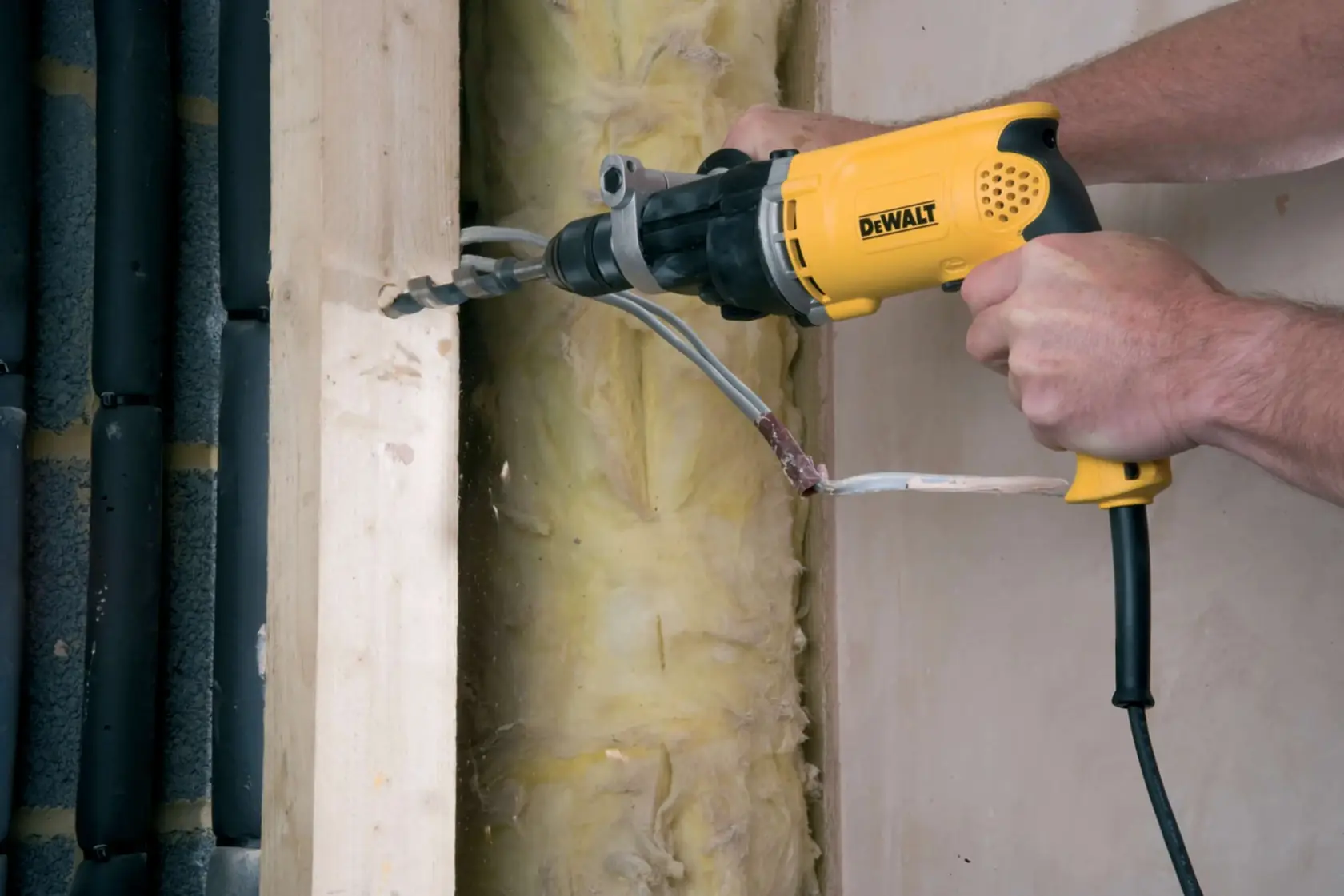 Une personne utilise une perceuse électrique jaune DEWALT pour percer un trou dans une poutre en bois, avec des câbles électriques et de l’isolant visibles à l’arrière-plan.