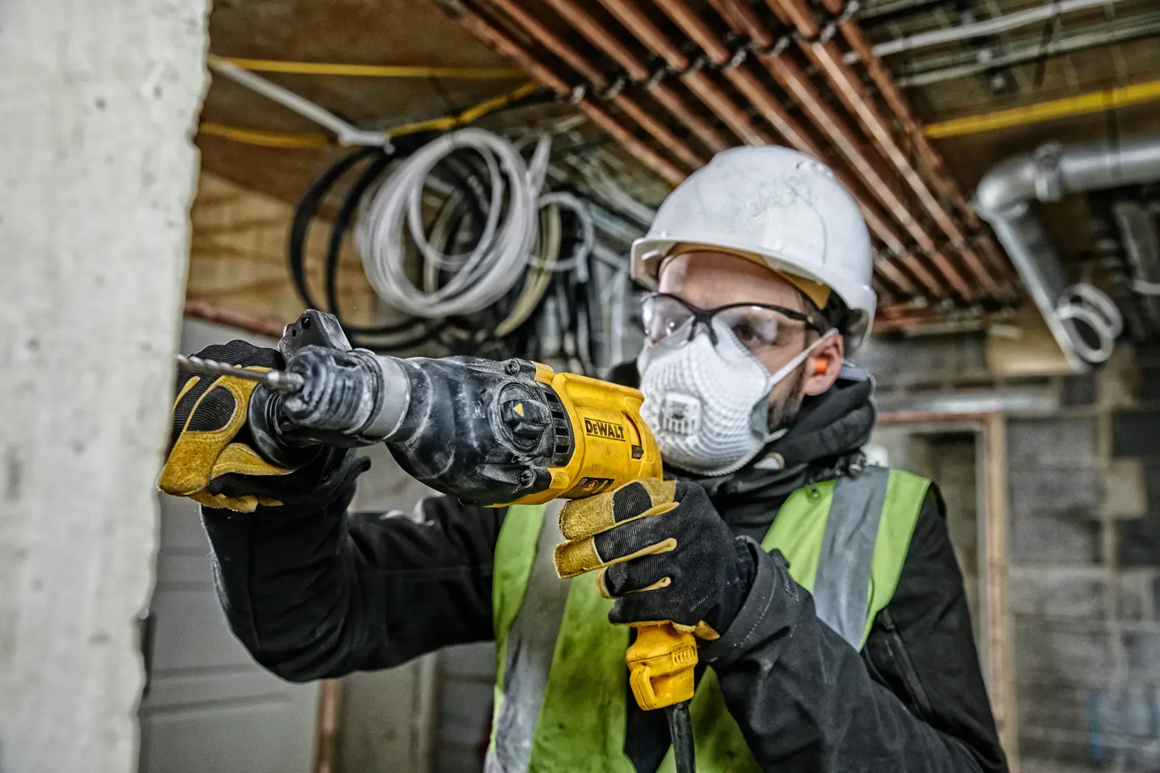 Un ouvrier du bâtiment portant un casque, des lunettes de protection, des gants et un gilet haute visibilité utilise une perceuse à percussion rotative DEWALT D25033 sur un chantier. En arrière-plan, on voit des murs non finis, des tuyaux et des câbles électriques.