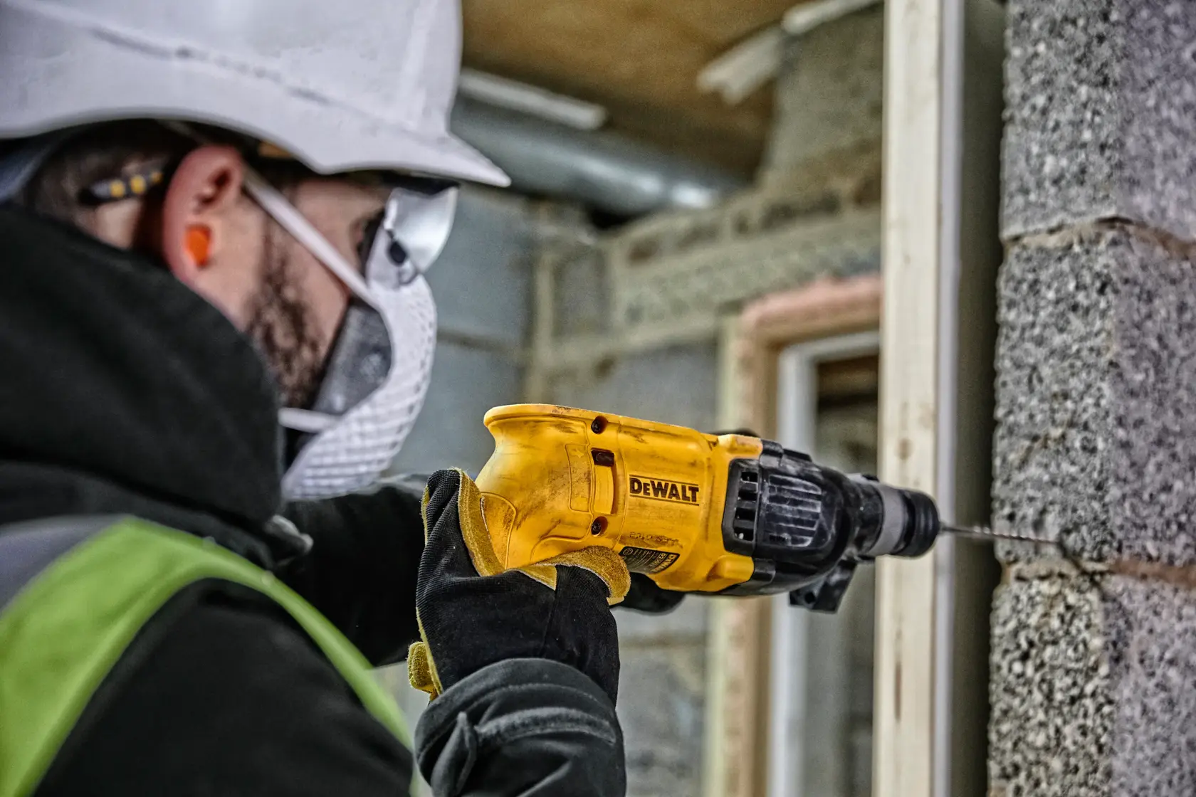 Une personne équipée de matériel de sécurité utilise une perceuse électrique jaune DEWALT pour percer un mur en béton sur un chantier.
