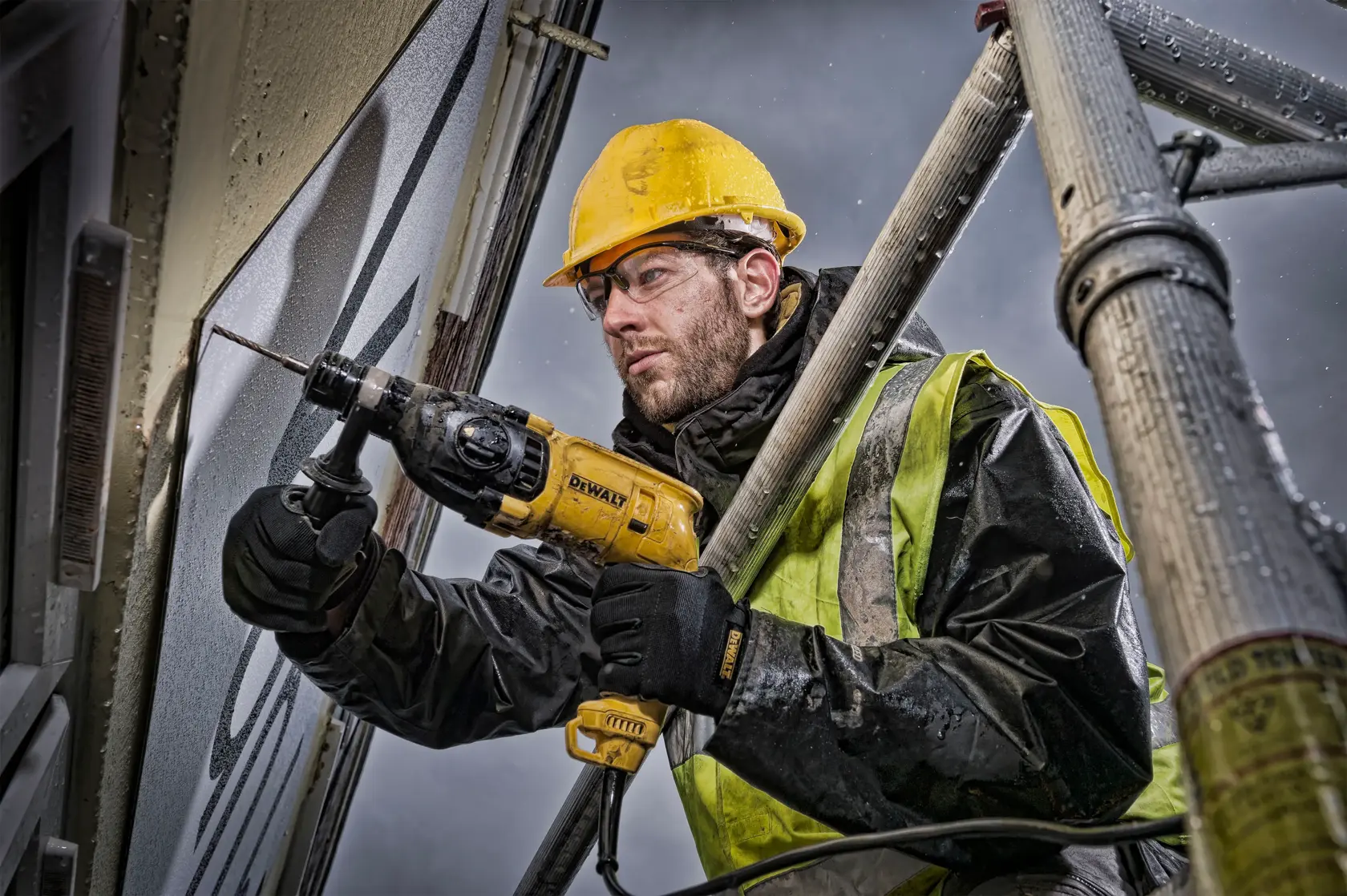 Un ouvrier de construction portant un casque de sécurité jaune et un gilet de sécurité utilise une perceuse à percussion DEWALT (modèle D25033) pour percer un mur depuis un échafaudage. Le visage de l'ouvrier est flouté pour préserver sa confidentialité.