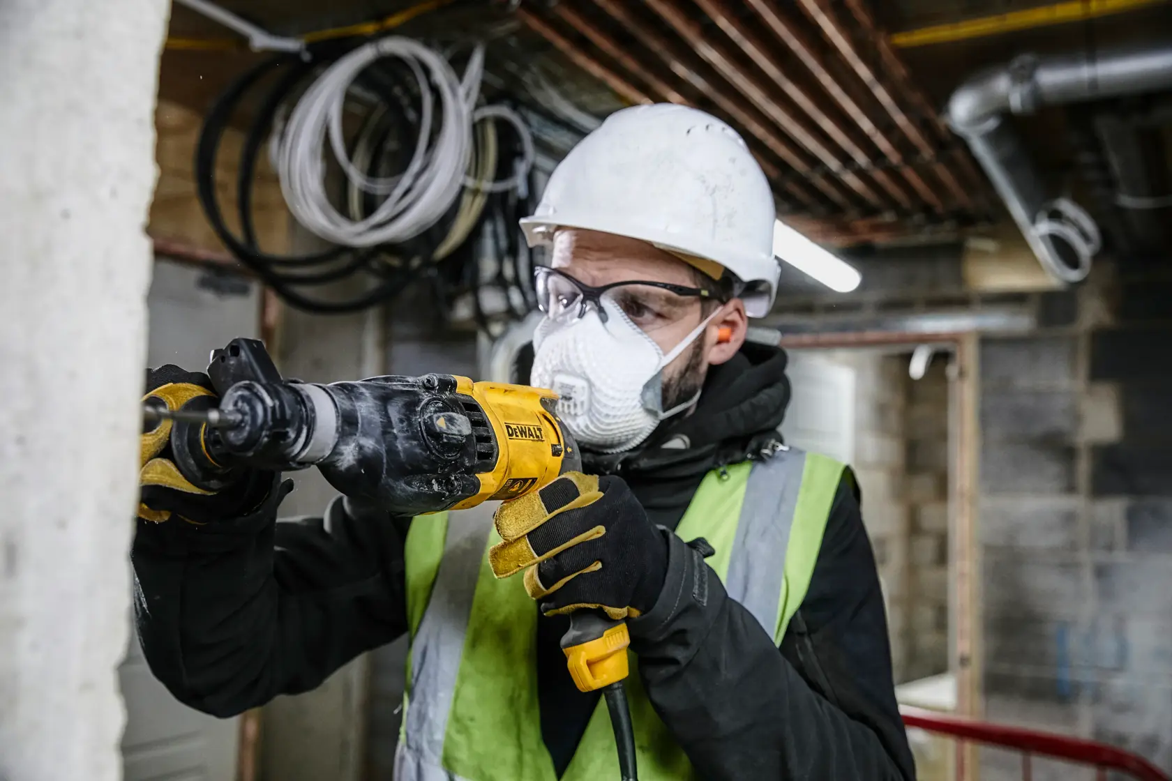 Eine Person mit Schutzhelm, Handschuhen und Warnweste benutzt eine DEWALT-Bohrmaschine, um an einer Betonwand in einer Bauumgebung zu arbeiten. Das Gesicht ist aus Datenschutzgründen verschwommen.