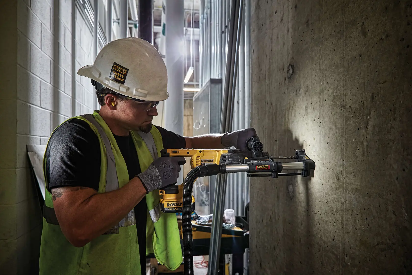 Un ouvrier du bâtiment portant un casque et un gilet de sécurité utilise un outil électrique jaune DEWALT avec un accessoire d'extraction de poussière pour percer dans un mur en béton dans un environnement industriel.