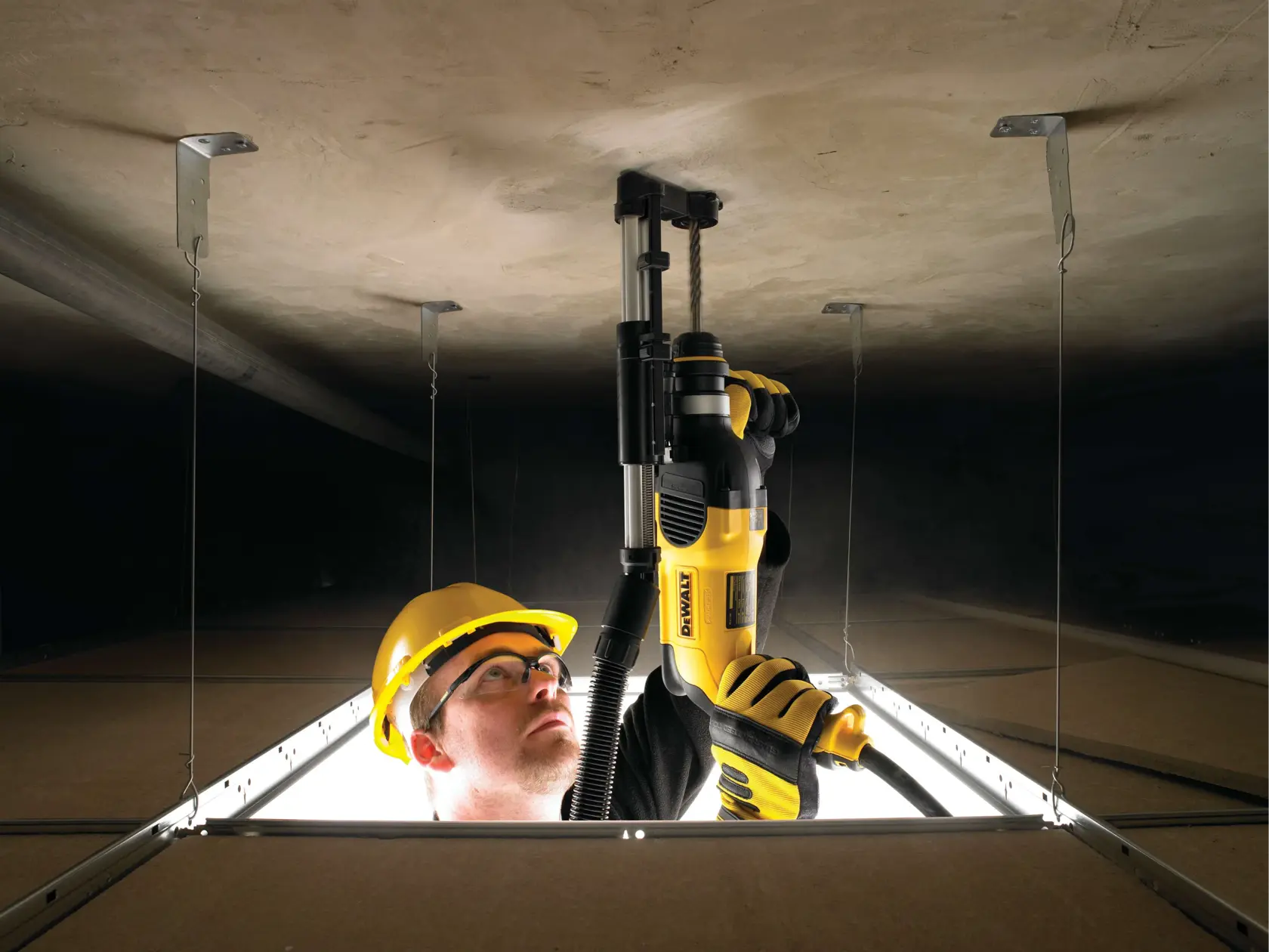 Une personne portant un équipement de sécurité et des gants utilise un outil électrique jaune DEWALT pour percer un plafond à l’intérieur d’un chantier, vue depuis une ouverture dans les dalles du plafond.