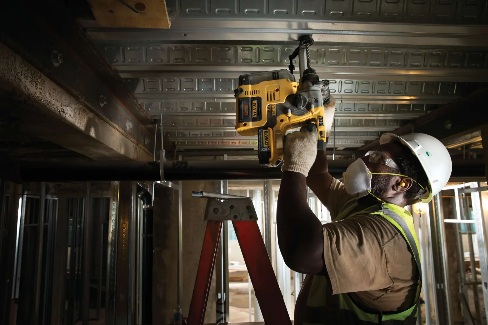 Un ouvrier du bâtiment portant un casque, un gilet de sécurité, des gants et un masque utilise un outil DEWALT D25303DH pour percer une poutre métallique au plafond tout en étant sur une échelle sur un chantier.