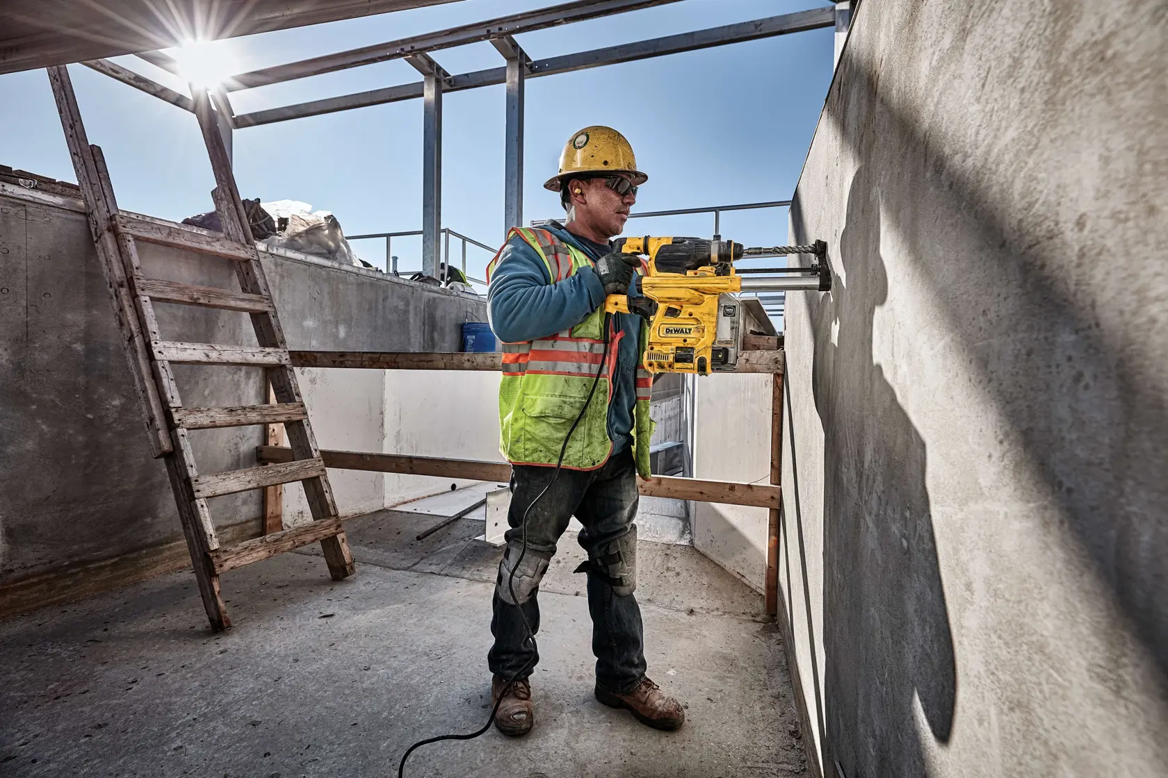 Un ouvrier du bâtiment portant un équipement de sécurité utilise une perceuse rotative DEWALT D25333K sur un mur en béton dans un chantier extérieur.