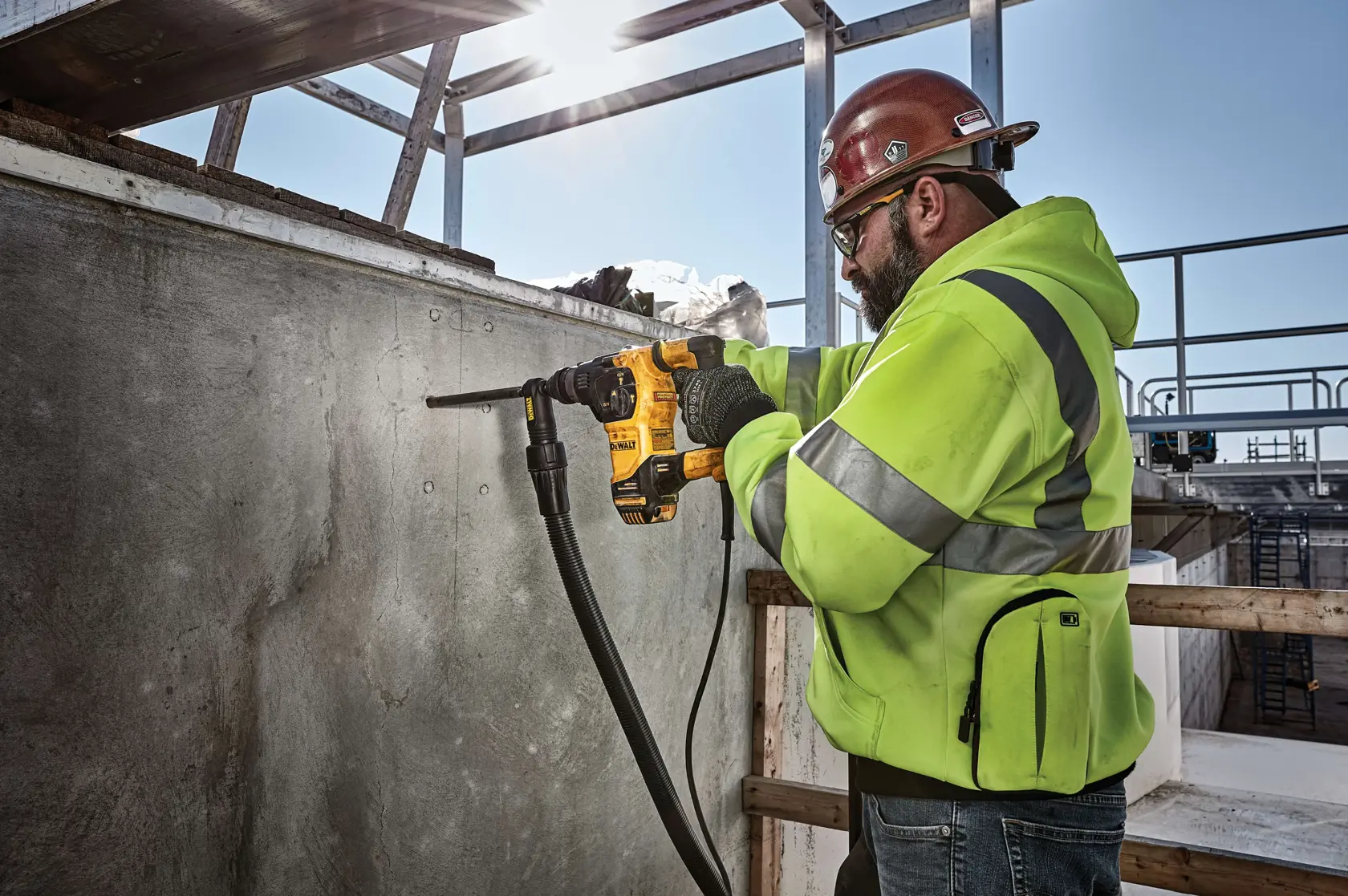 Un ouvrier du bâtiment portant un gilet de sécurité et un casque utilise un marteau perforateur rotatif DEWALT D25333K pour percer un mur en béton sur un chantier. Le visage de l'ouvrier est flouté pour préserver sa vie privée.