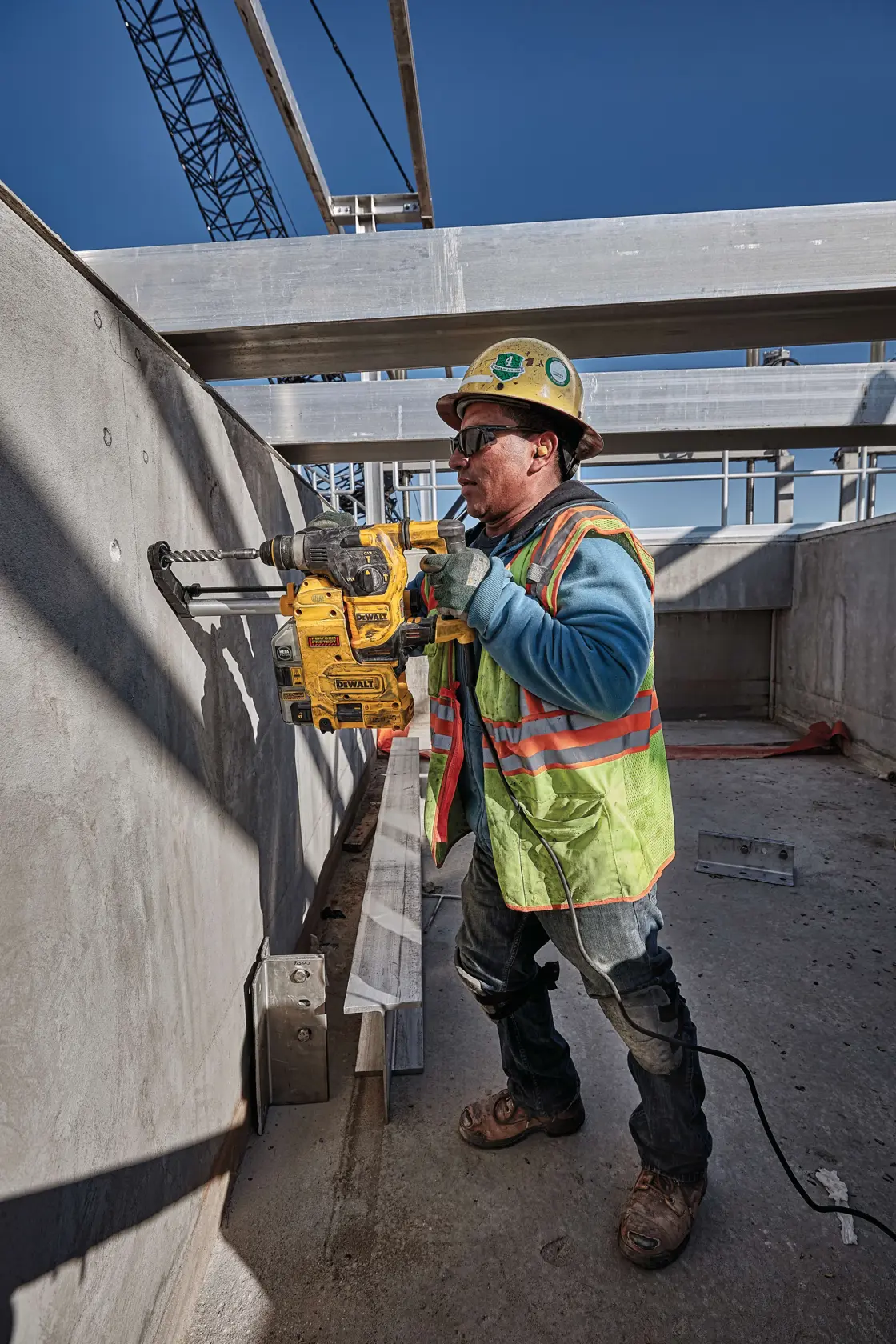 A construction worker wearing a safety helmet and high-visibility vest operates a yellow DEWALT rotary hammer drill, model D25333K, to bore a hole into a concrete wall at an outdoor construction site. The worker stands near a wooden ladder under a partly built structure with sunlight streaming in.