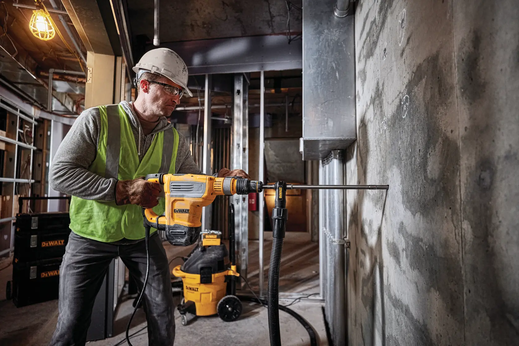 Un ouvrier du bâtiment portant un casque jaune et un gilet de sécurité utilise une perceuse à percussion rotative DEWALT (modèle D25614K) pour percer un mur en briques, avec de la poussière expulsée par un système d'extraction de poussière attaché.