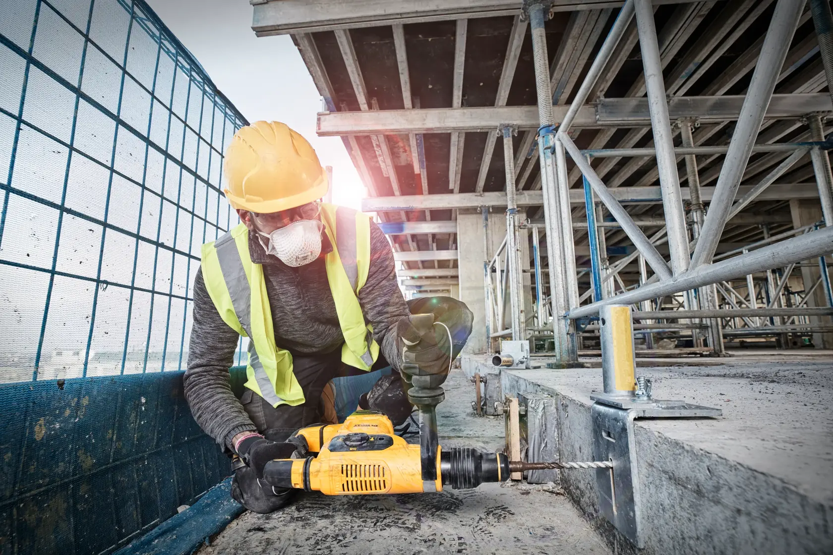 Un ouvrier du bâtiment portant un casque de sécurité jaune et un gilet réfléchissant utilise une perceuse à percussion jaune DEWALT pour percer une surface en béton sur un chantier.