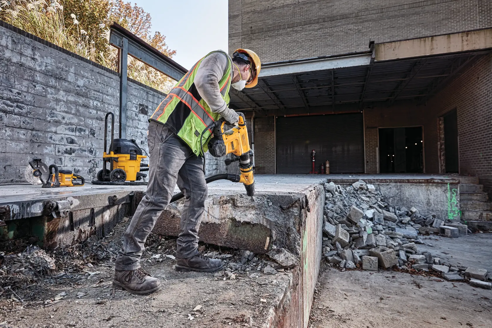 Un ouvrier de chantier portant des équipements de sécurité utilise un marteau démolisseur DEWALT sur une structure en béton sur un chantier. Du béton effondré et des débris sont visibles autour de la zone.