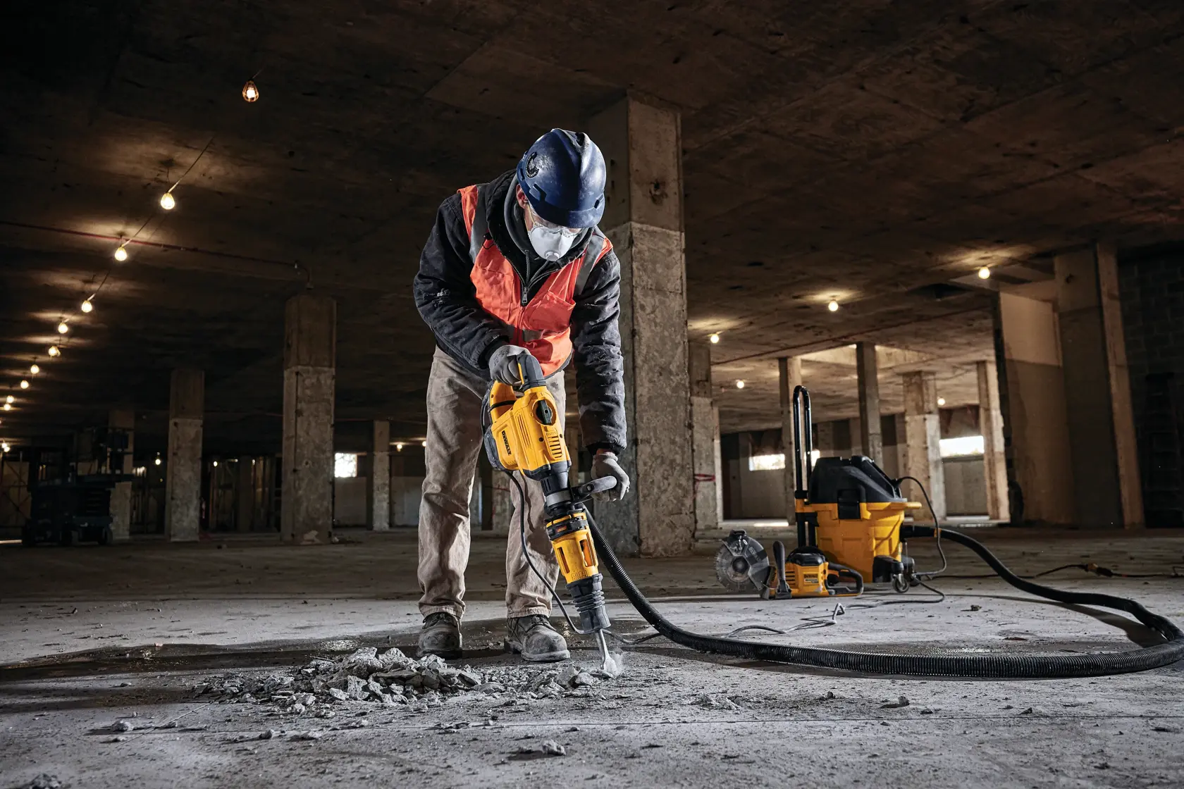 Une personne portant un équipement de sécurité, dont un casque et un masque facial, utilise un marteau de démolition DEWALT jaune pour casser du béton sur le sol d'un grand chantier de construction. Une machine d'aspiration DEWALT jaune est visible à l'arrière-plan.
