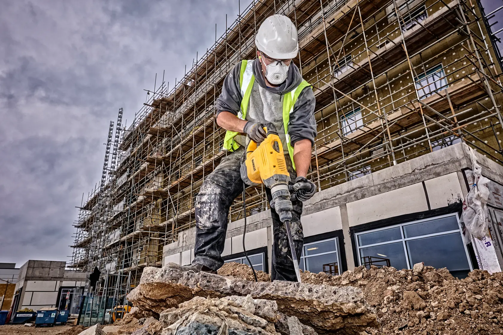 Un ouvrier du bâtiment portant un casque de sécurité et un gilet haute visibilité utilise un marteau de démolition jaune DEWALT pour casser du béton sur un chantier. Des échafaudages et un bâtiment en cours de construction sont visibles en arrière-plan.