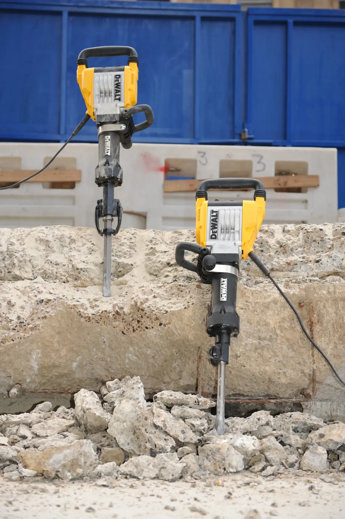 Deux marteaux-piqueurs électriques DEWALT sont montrés en train de casser une grande dalle de béton, avec des gravats et des morceaux de béton brisés au sol. Les outils sont positionnés verticalement et reliés par des câbles, dans un environnement de chantier extérieur.