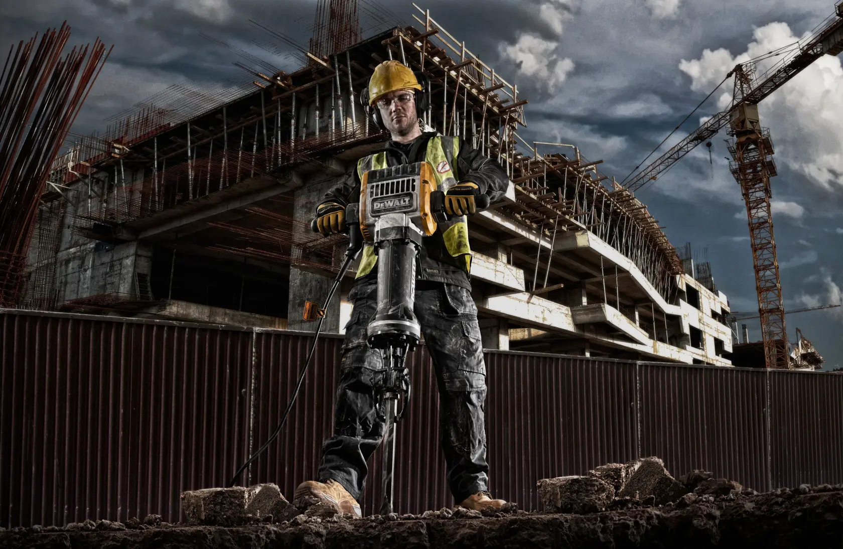 Un ouvrier de construction utilisant un marteau de démolition DEWALT D25981 sur un chantier, avec une structure partiellement construite et des grues en arrière-plan.
