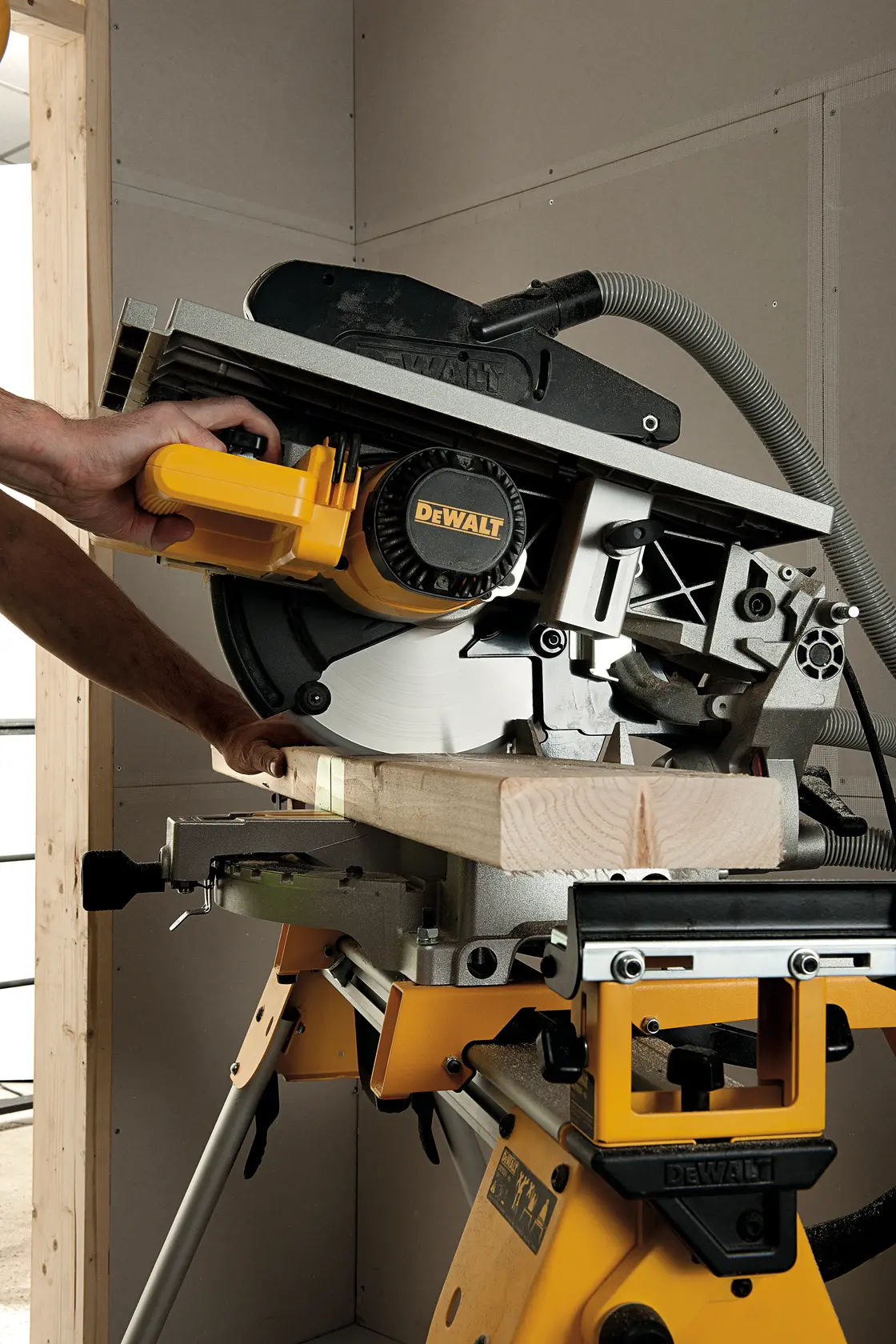 Une personne utilise une scie DEWALT D27113 pour couper une pièce de bois. La scie est montée sur un support jaune et fonctionne dans un atelier intérieur.