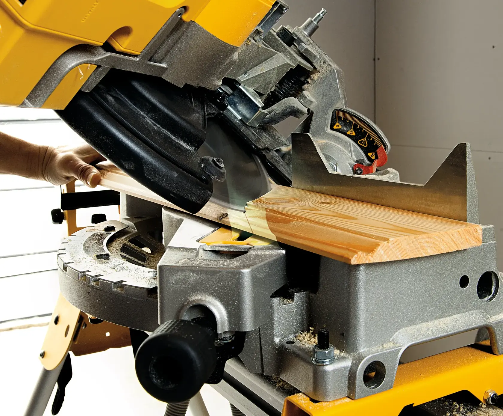 Gros plan sur une scie à onglet DEWALT coupant une pièce de bois en angle. La scie est montée sur un établi et une main tient la pièce de bois en place. De la sciure est visible sur la surface.