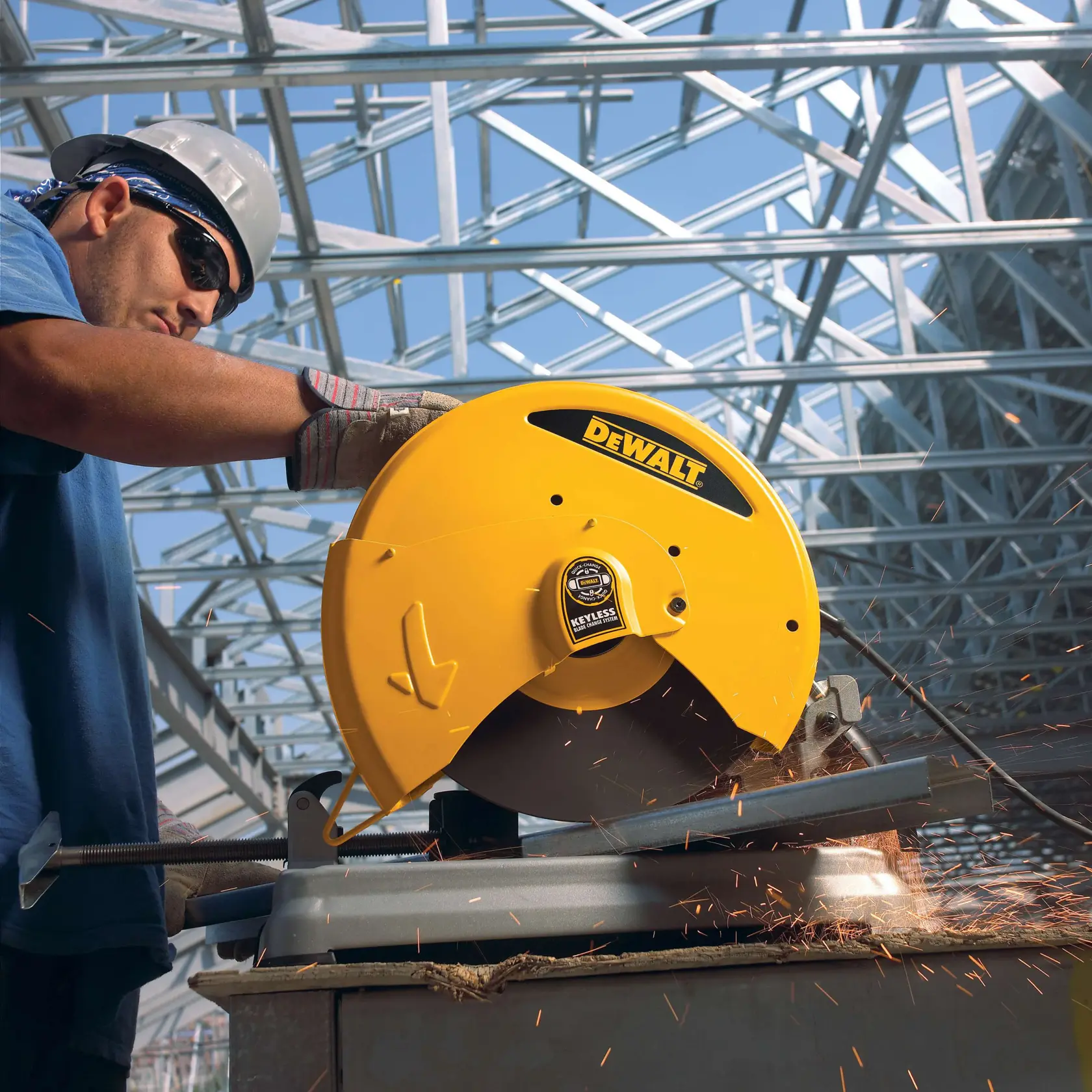 Une personne portant des gants de travail et un casque opère une scie à métaux jaune DEWALT pour couper du métal, avec des étincelles, à l'intérieur d'un bâtiment avec une structure en acier.