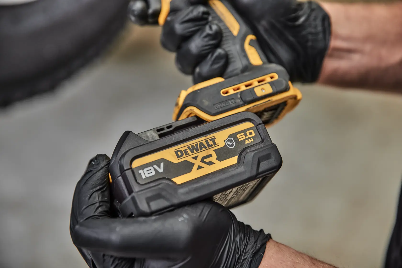 Vue en gros plan d'une personne portant des gants noirs tenant un outil électrique sans fil DEWALT avec une batterie lithium-ion XR 18V DCB184G attachée.