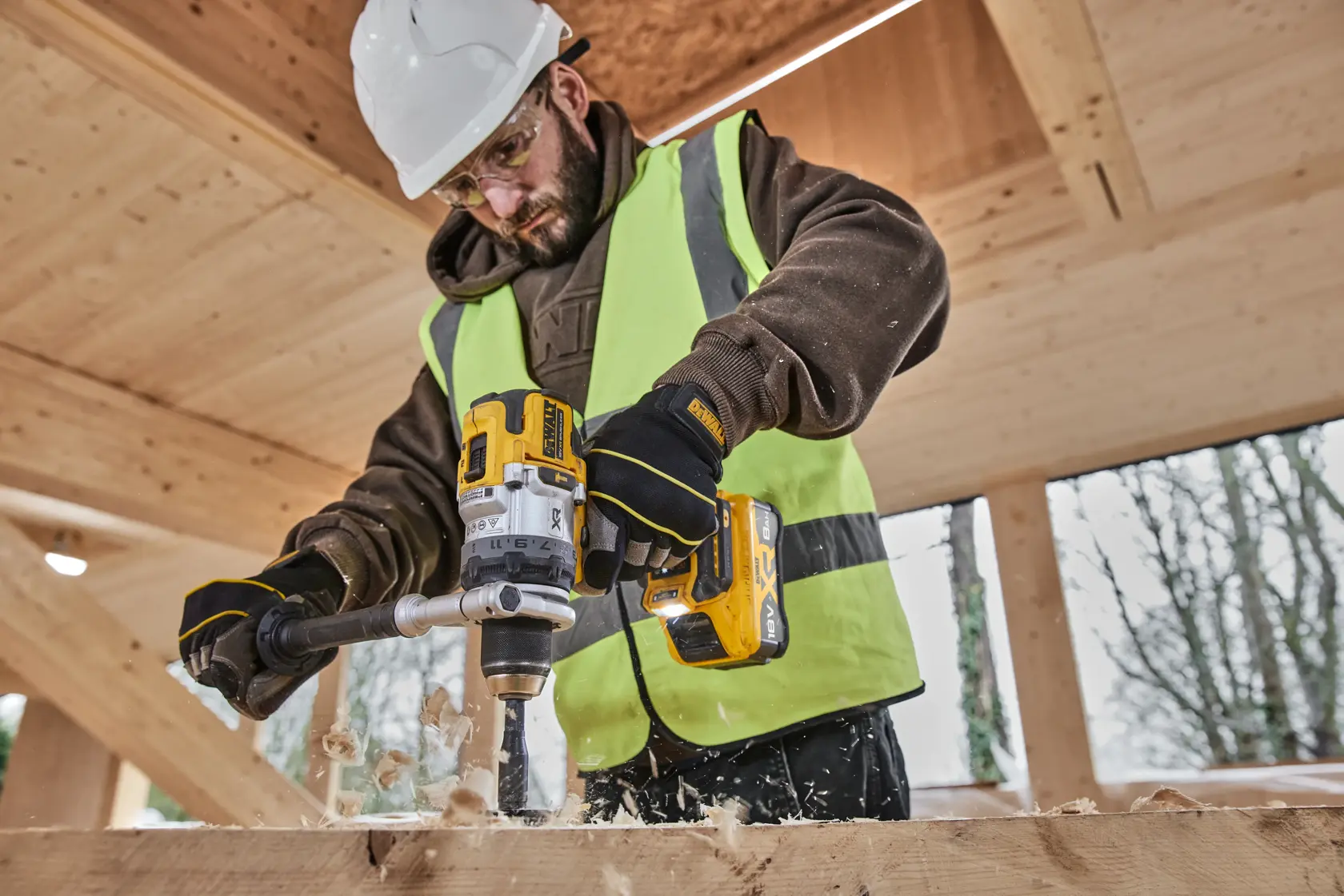 Person bohrt Holz mit DEWALT DCB1880. Trägt Sicherheitsweste, Handschuhe und Helm auf Baustelle.