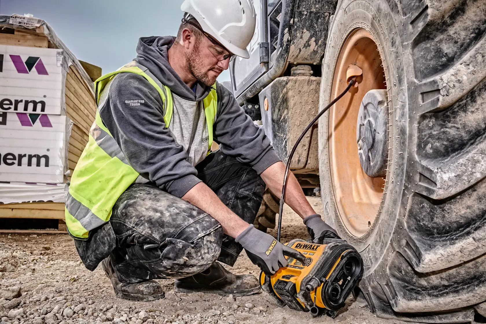 Eine Person mit Sicherheitshelm und Warnweste verwendet einen DEWALT DCC018N Akku-Kompressor, um den Reifen eines großen Baustellenfahrzeugs auf einer Baustelle aufzupumpen.