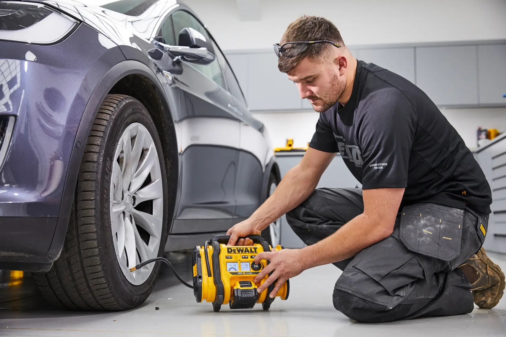 Eine Person kniet auf dem Boden und benutzt einen tragbaren DEWALT DCC018N Luftkompressor, um den Reifen eines geparkten Autos in einer Werkstatt oder Garage aufzupumpen. Das Gesicht der Person ist aus Datenschutzgründen unscharf.