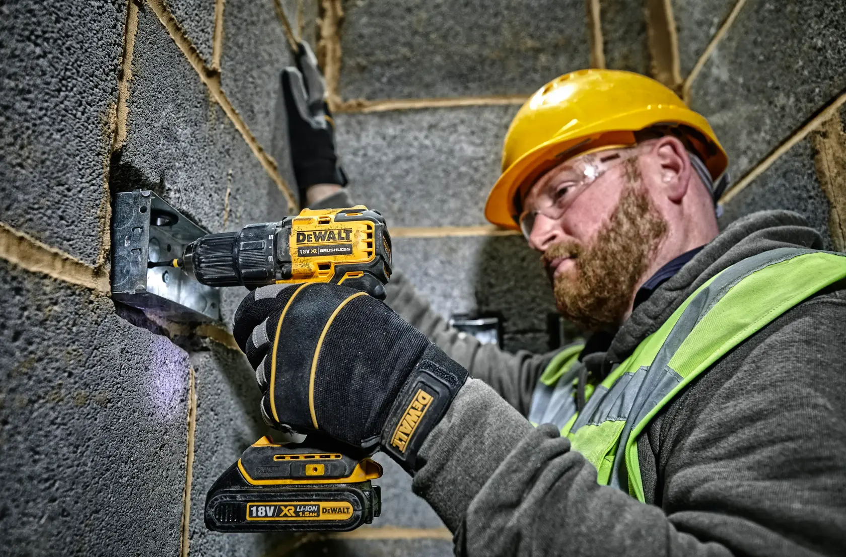 Un ouvrier du bâtiment portant un casque de sécurité jaune et des gants utilise une perceuse sans fil DEWALT DCD708 pour fixer une équerre métallique à un mur en blocs de béton. Le visage de l'ouvrier est flouté, et il porte un gilet haute visibilité ainsi qu'un sweat à capuche gris.