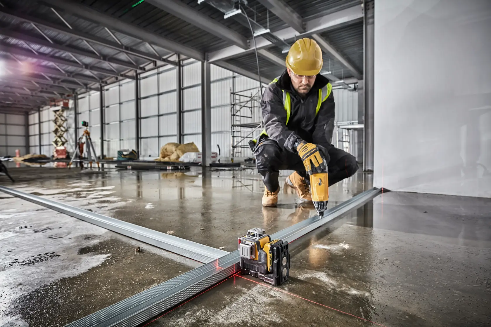 Un ouvrier du bâtiment utilise un niveau laser Dewalt (DCE089D1R) pour aligner des cadres en métal sur un sol en béton humide dans un environnement industriel. L'ouvrier porte un casque de sécurité jaune et utilise une perceuse électrique en étant à genoux.