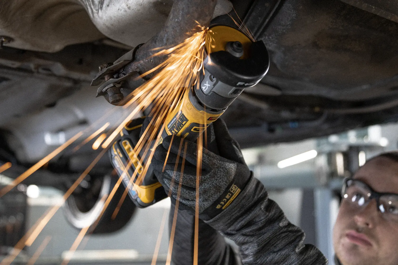 Meuleuse DEWALT coupant du métal sous un véhicule, avec des étincelles. Gants de protection portés.