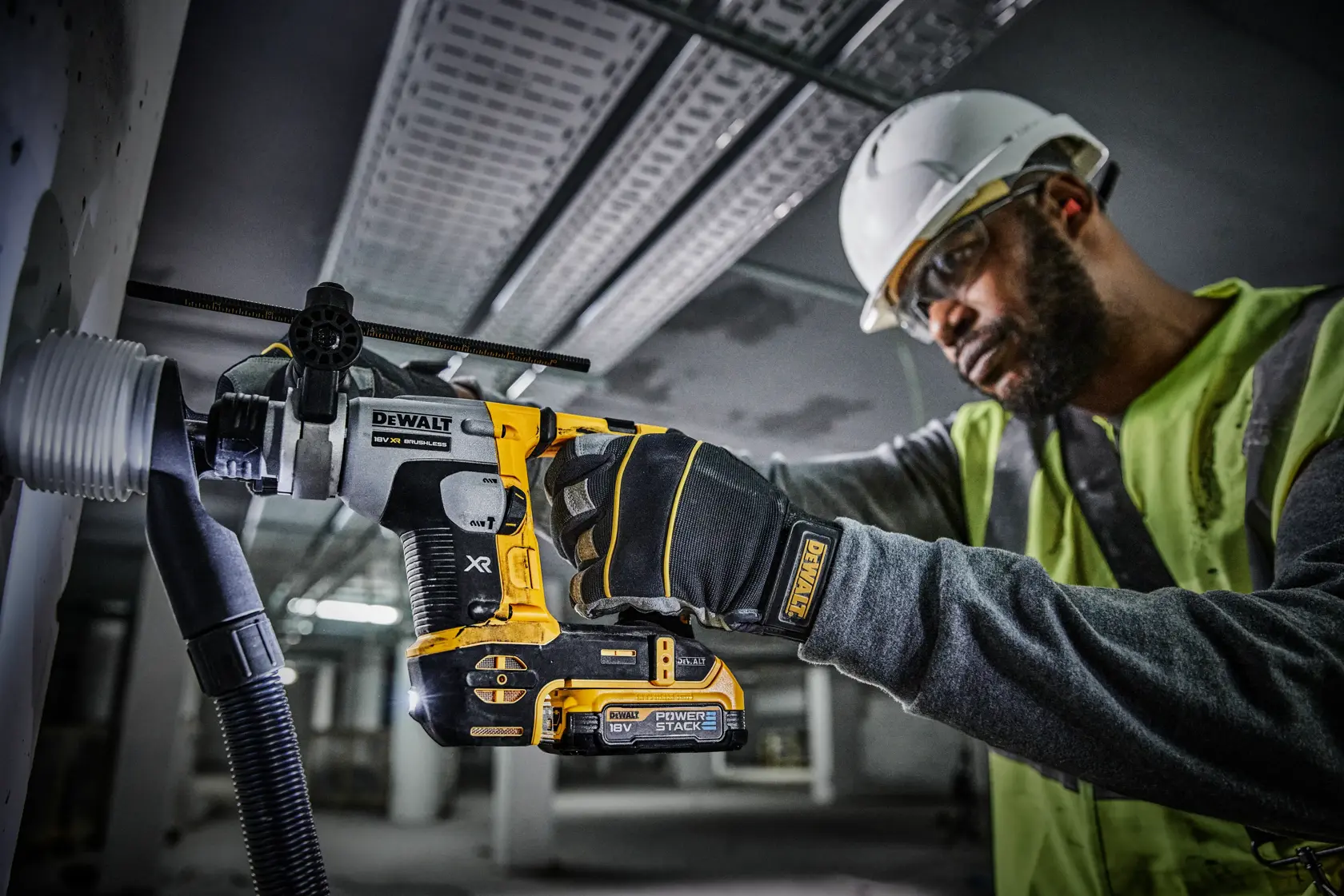 Une personne portant un casque de sécurité et un gilet haute visibilité utilise une perceuse rotative sans fil DEWALT pour percer un mur, avec un équipement d'extraction de poussière attaché. La perceuse porte les inscriptions XR et Power Stack.