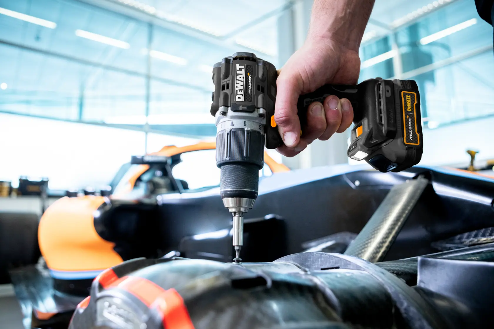 Gros plan sur une personne utilisant une perceuse sans fil DEWALT pour fixer ou assembler des pièces sur ce qui semble être un composant de véhicule, avec une voiture orange visible en arrière-plan.