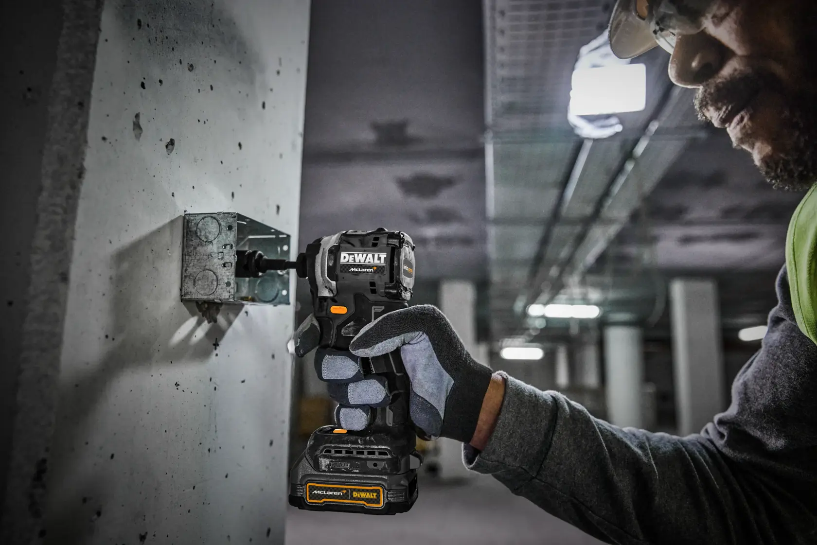 Une personne portant des gants de sécurité utilise une perceuse sans fil DEWALT pour fixer une pièce métallique à un mur en béton dans un environnement industriel.