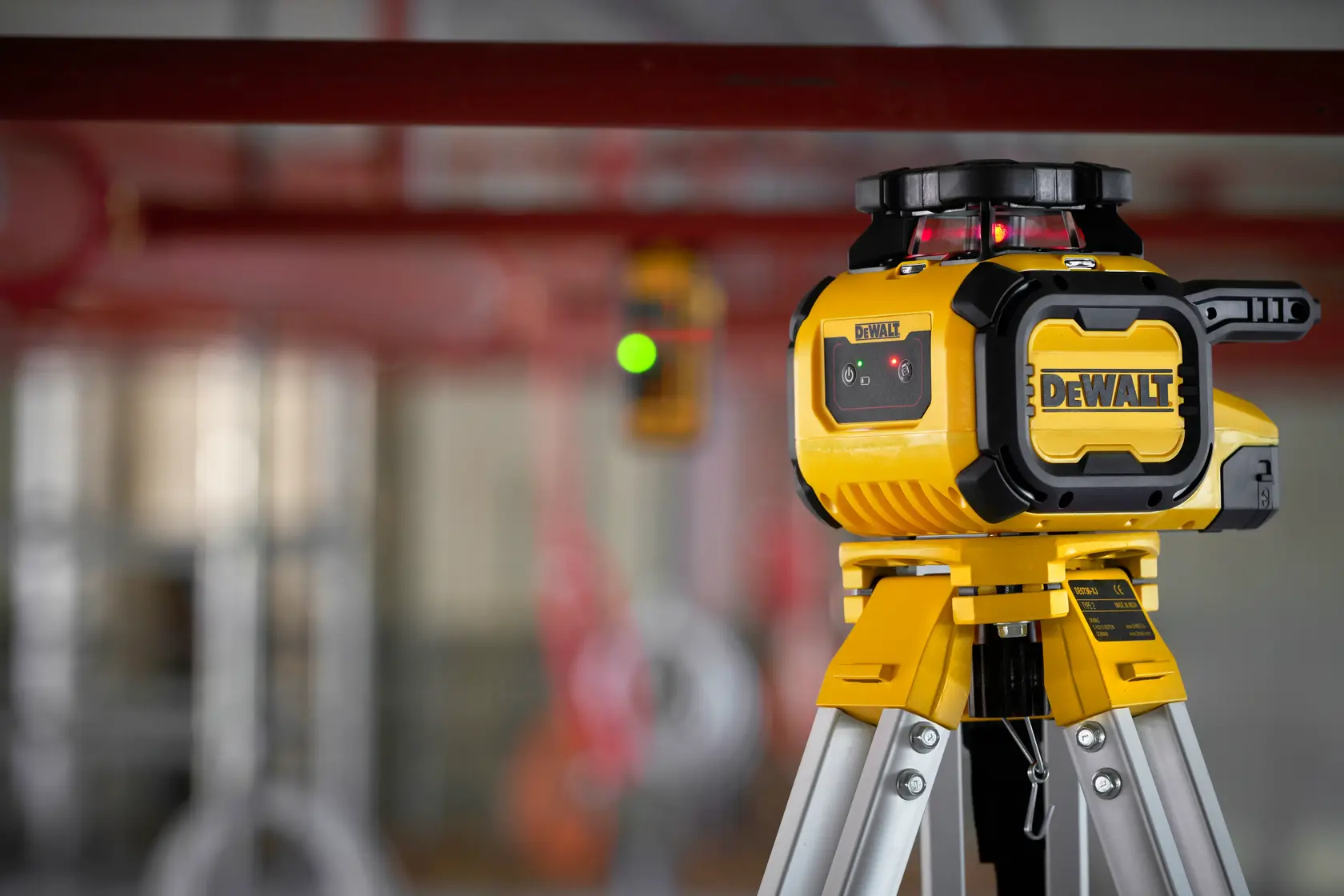 Niveau laser DEWALT DCLE05211R sur trépied dans un espace industriel, orienté vers des tuyaux rouges.