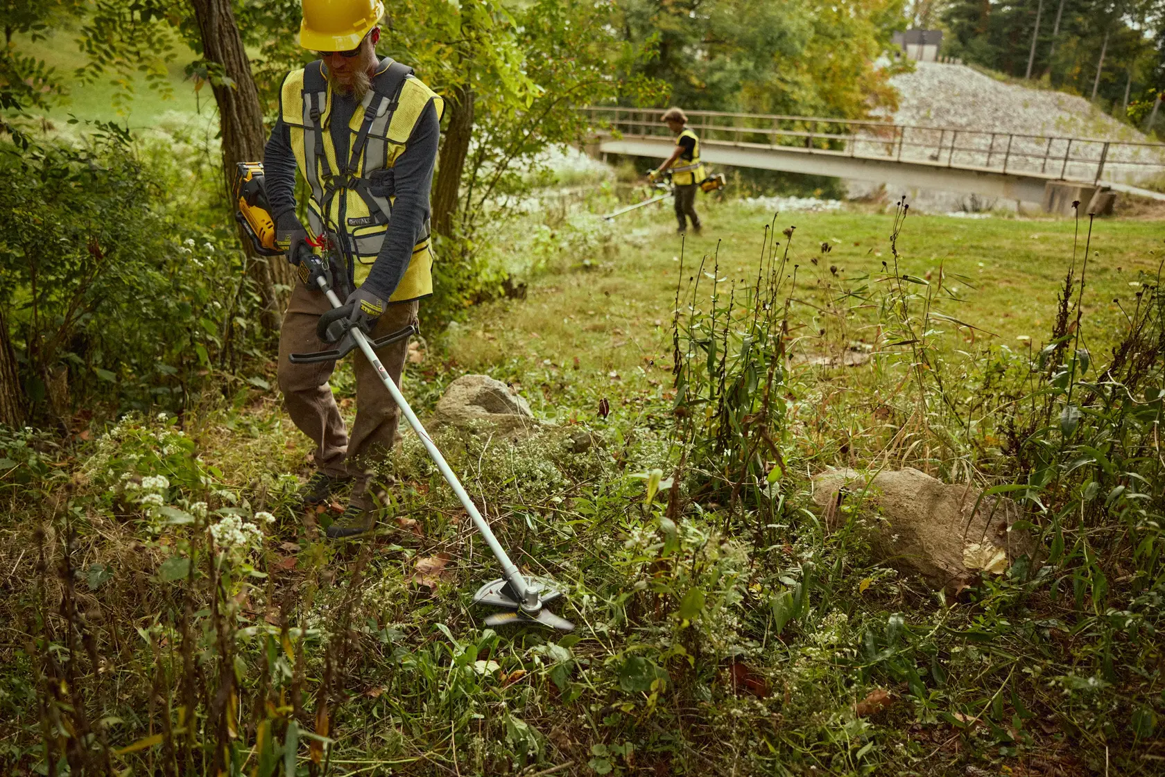 Deux personnes utilisent les coupe-bordures DEWALT DCMBC812N-XJ et DCMBC812N pour tailler l’herbe près des arbres et d’un