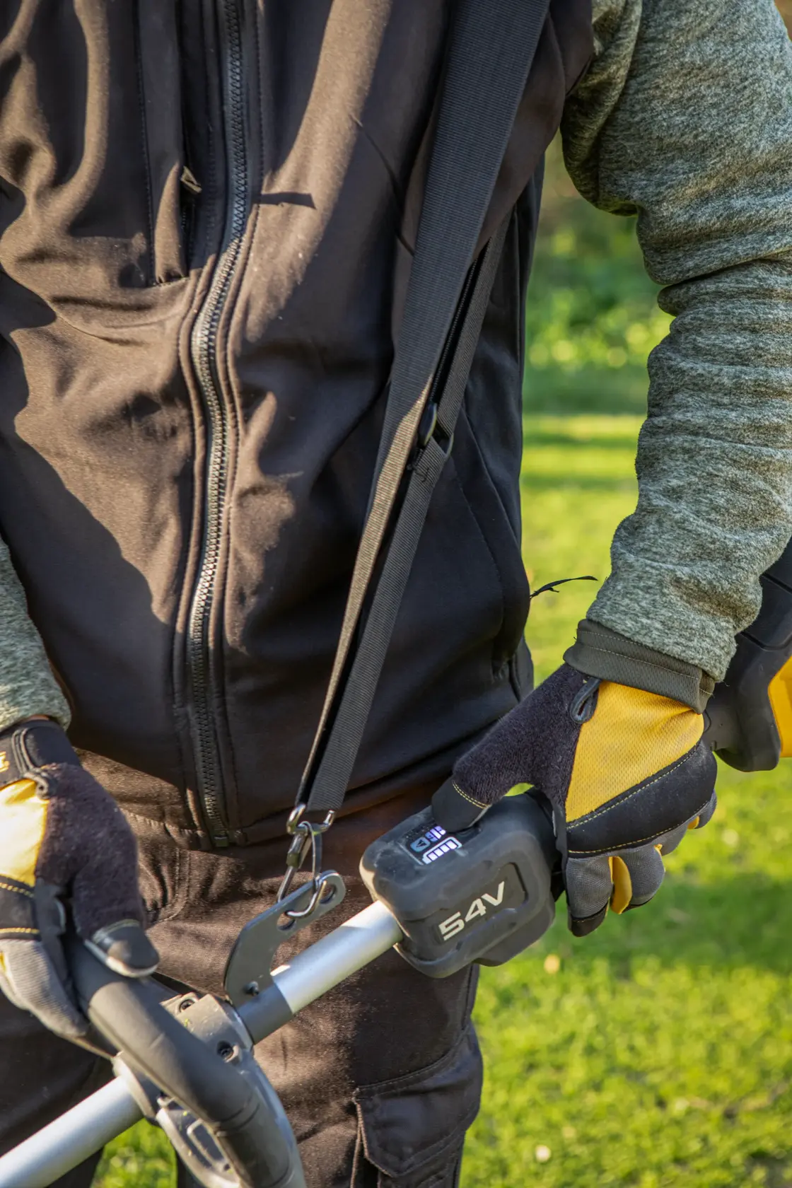 Personne avec des gants tenant un outil de jardin DEWALT 54V dehors, portant une veste à fermeture éclair.