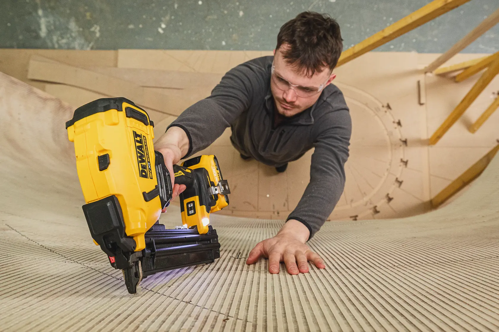 Personne utilisant une cloueuse DEWALT sur une surface en bois courbée, lunettes de sécurité, haut gris.