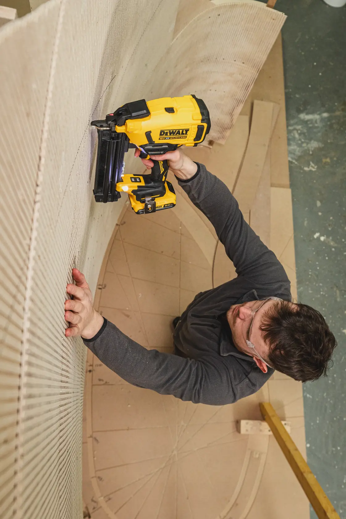 Personne utilise une cloueuse DEWALT DCN210SN-XJ sur bois courbé en atelier, lunettes de sécurité et vêtements foncés.