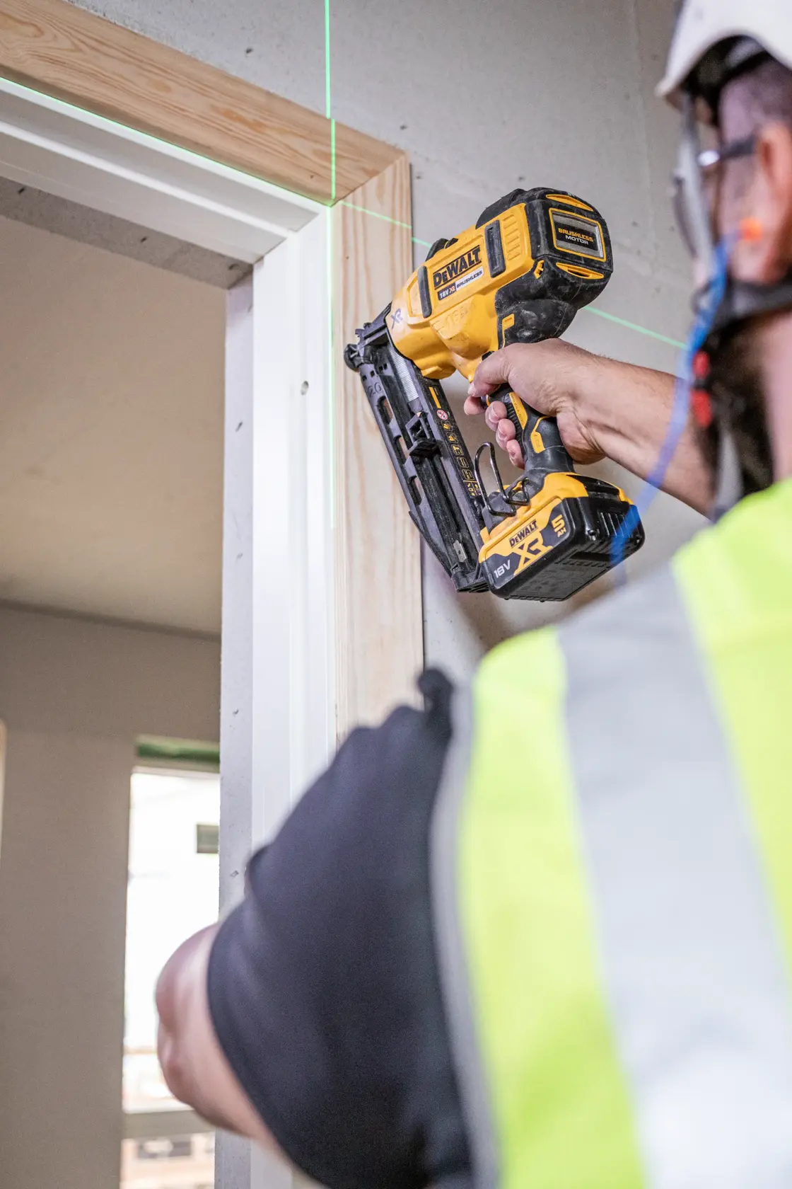 Person uses a DEWALT nail gun to install a wooden frame around a doorway in a construction setting.