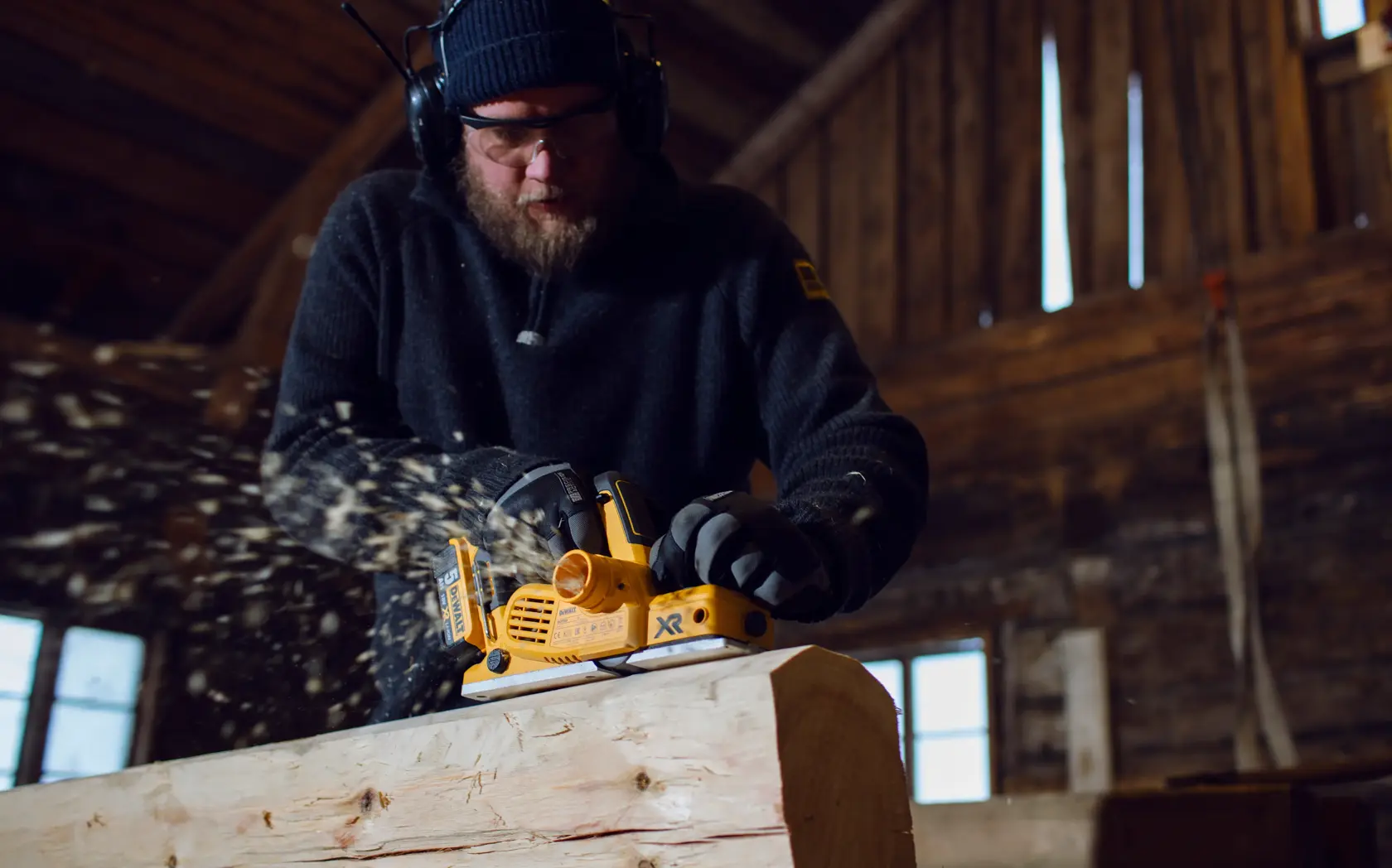 Une personne utilise un rabot sans fil DEWALT DCP580P2 pour aplanir une pièce de bois dans un atelier, des copeaux de bois s’envolent. Le visage de la personne est flouté et elle porte un casque de protection ainsi que des gants.