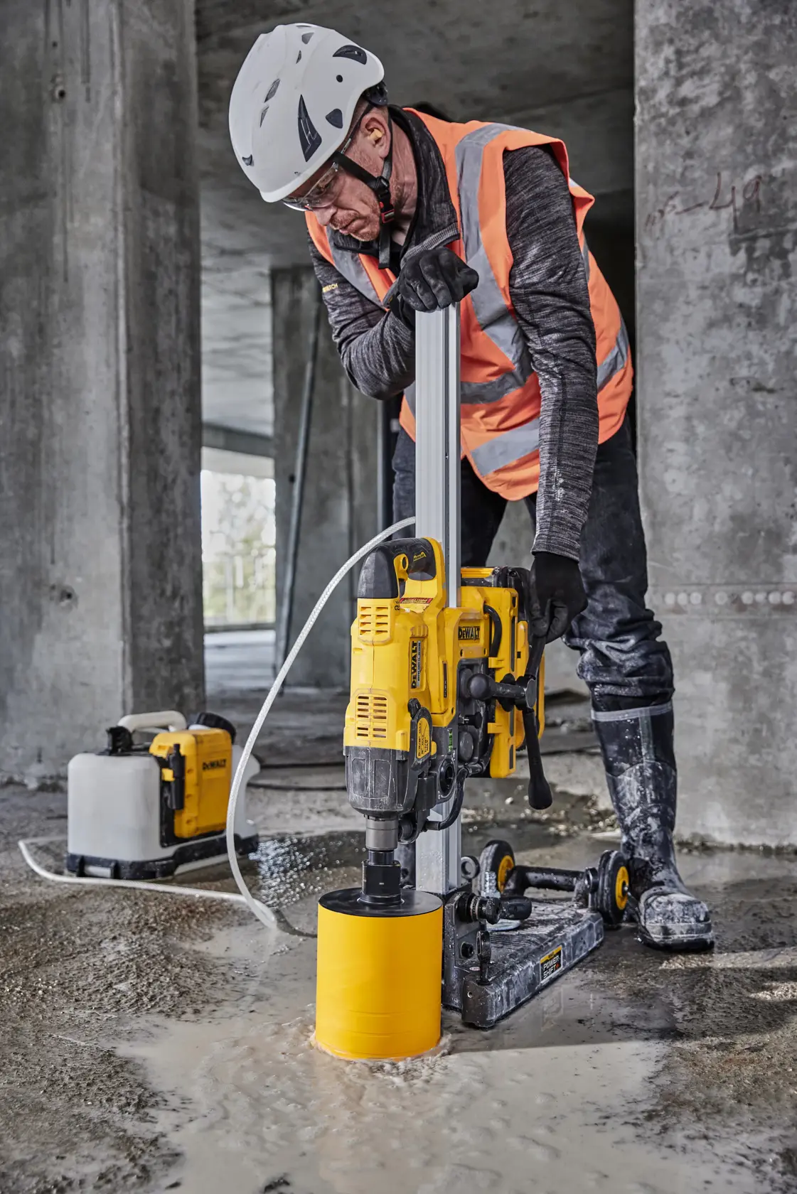 Un outil électrique DEWALT avec une base de vide intégrée est installé par un travailleur portant des gants et un casque de sécurité sur une surface en béton. L'outil comporte des roues jaunes et est connecté à un tuyau. Le texte sur l'image indique : « Base de vide intégrée pour une installation rapide et sécurisée. »