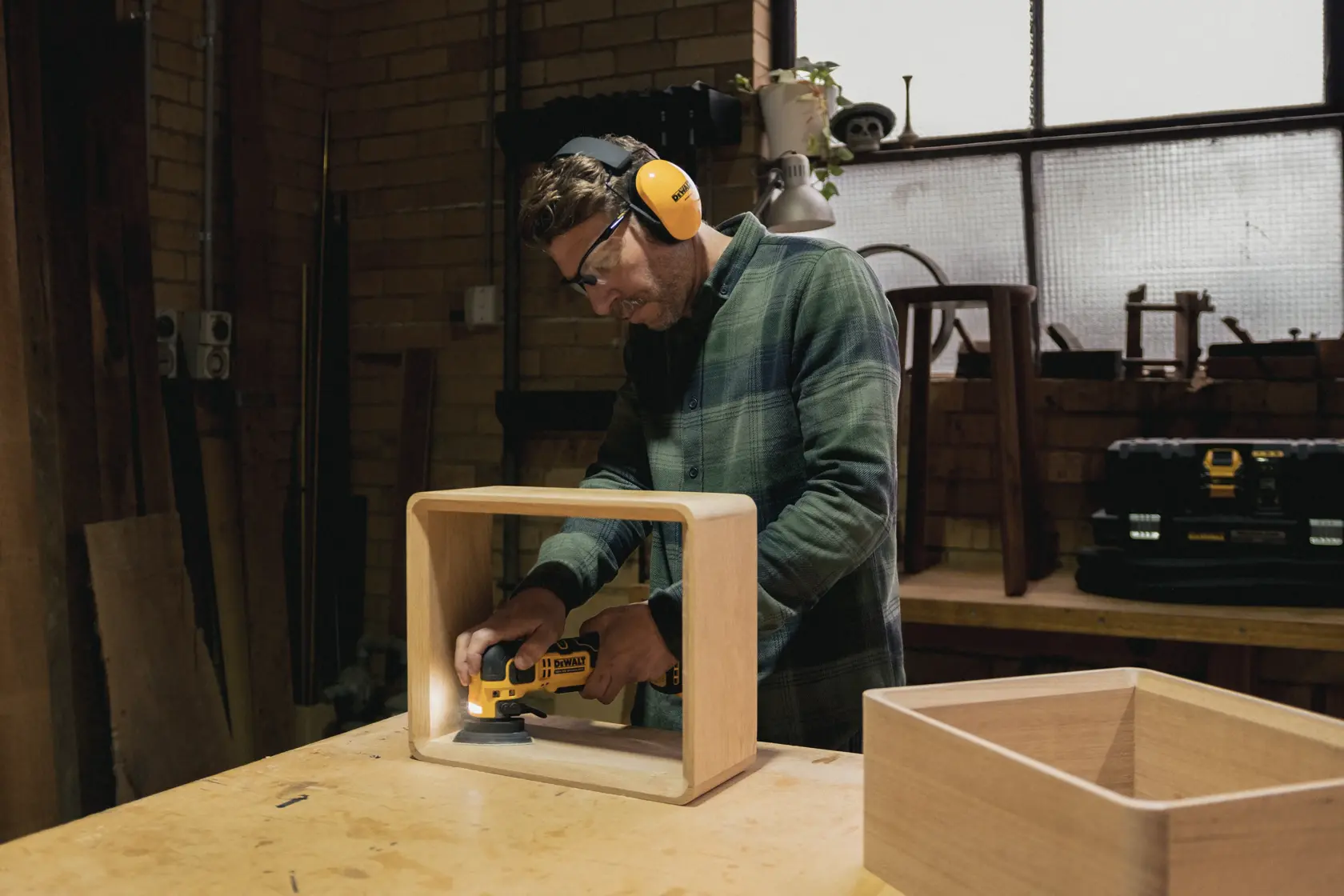 Personne ponçant une boîte en bois avec un outil DEWALT dans un atelier, portant un équipement de sécurité.