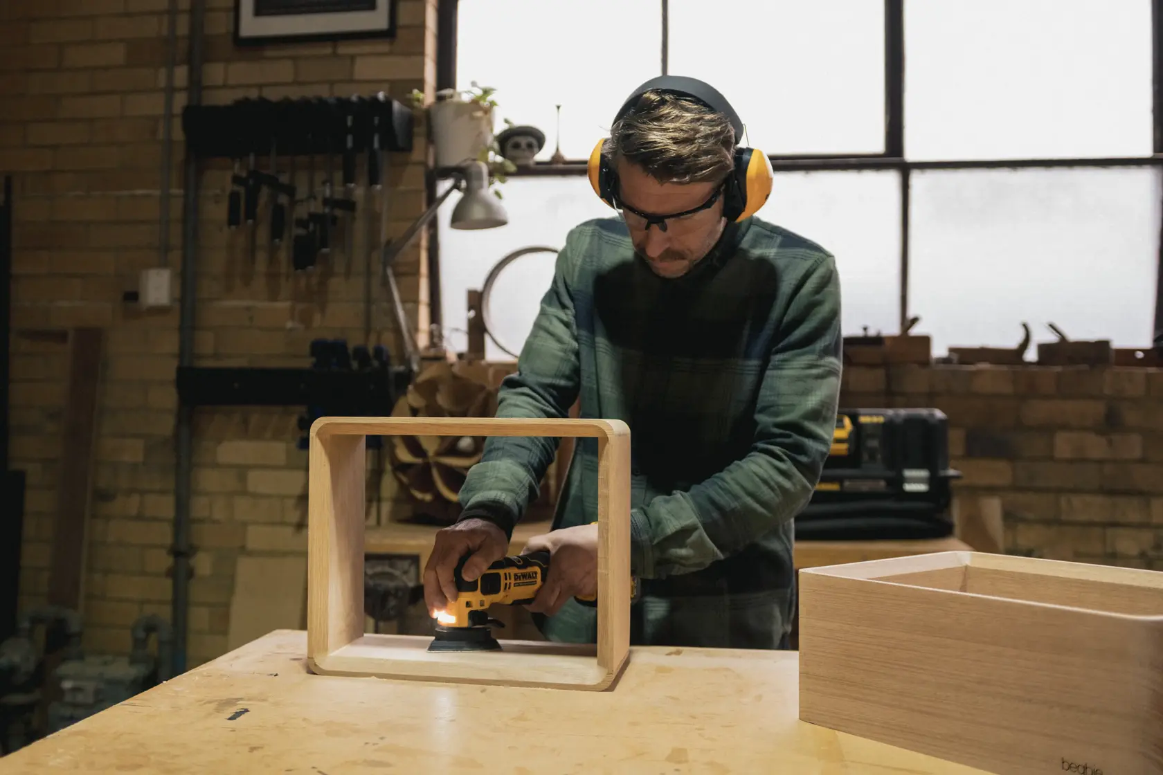 Personne ponçant un cadre en bois avec une ponceuse DEWALT, portant casque et lunettes en atelier.