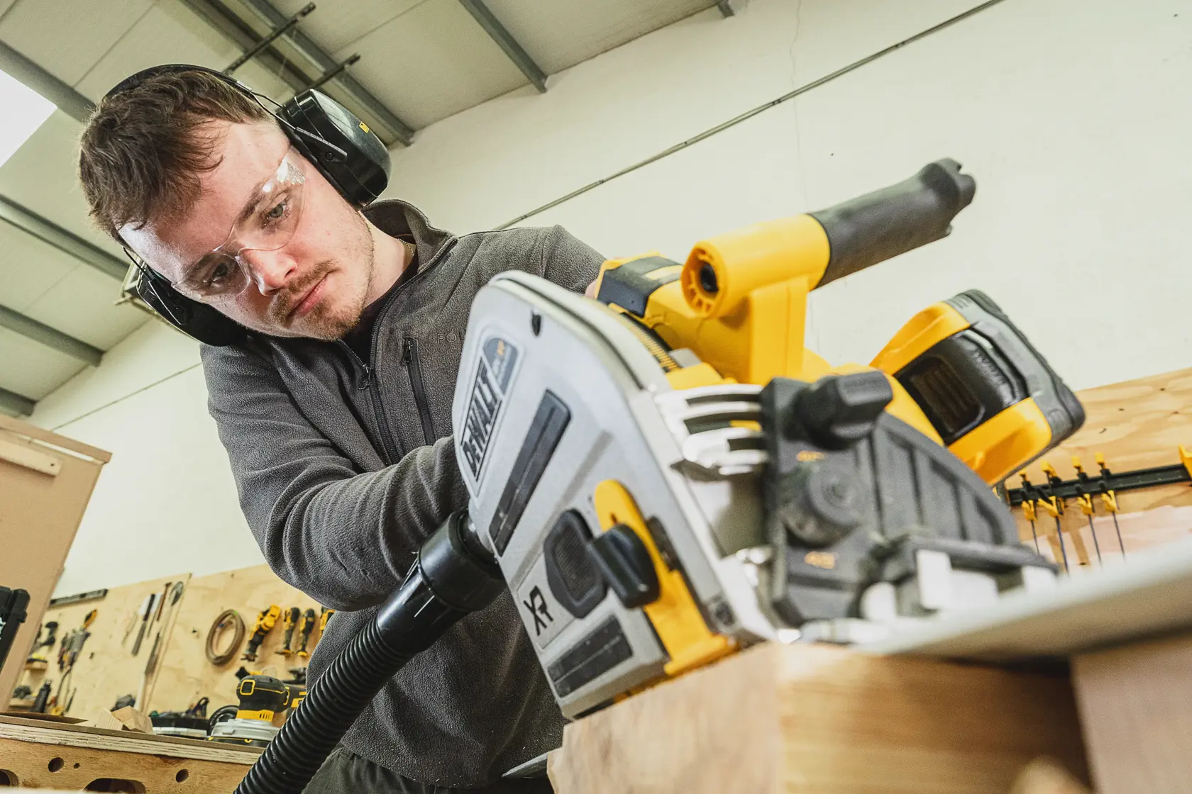 Person nutzt DEWALT Akku-Kreissäge an Holz, trägt Schutzbrille und Gehörschutz in einer Werkstatt.