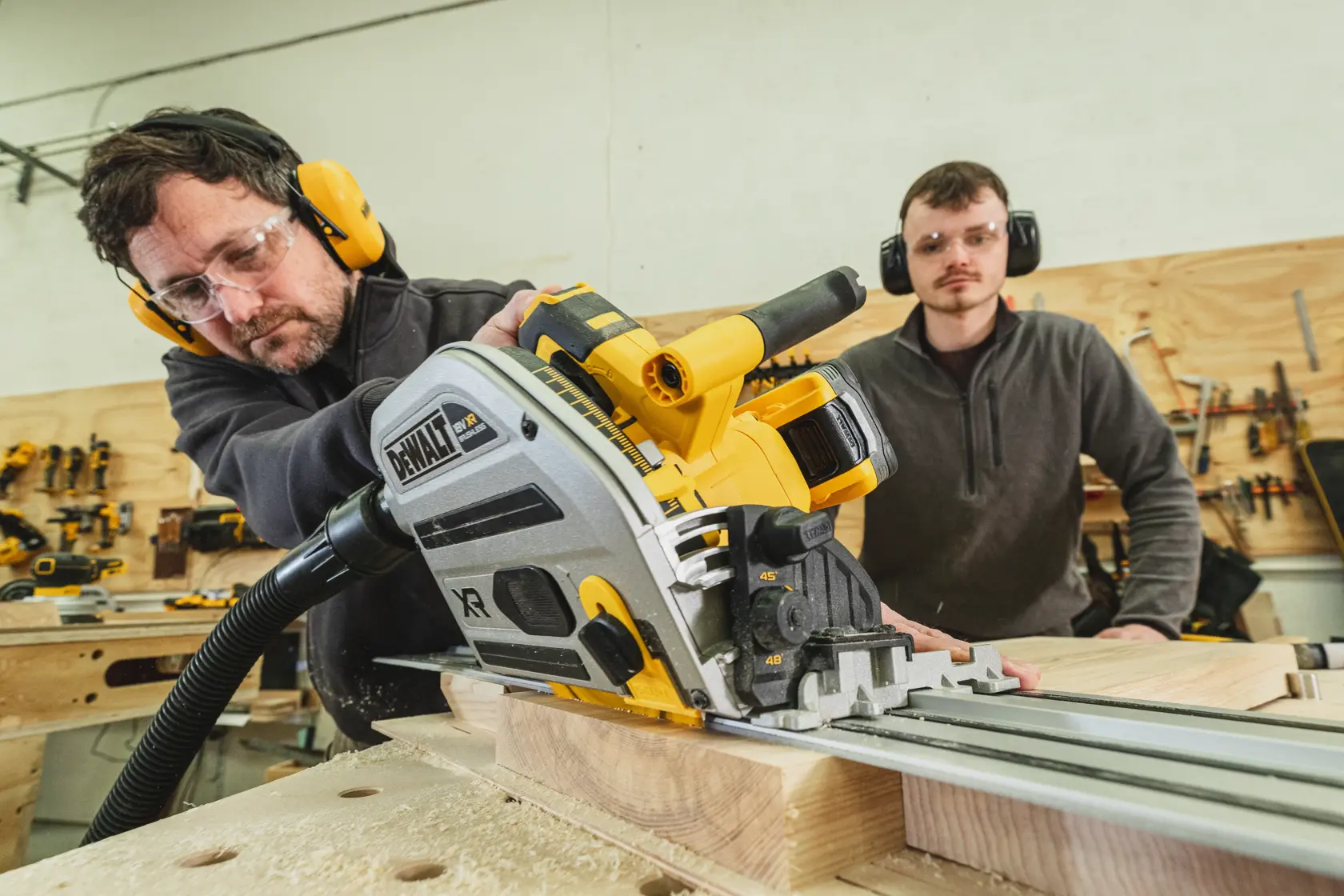 Mann sägt Holz mit einer DEWALT Führungssäge, eine weitere Person beobachtet in der Werkstatt.
