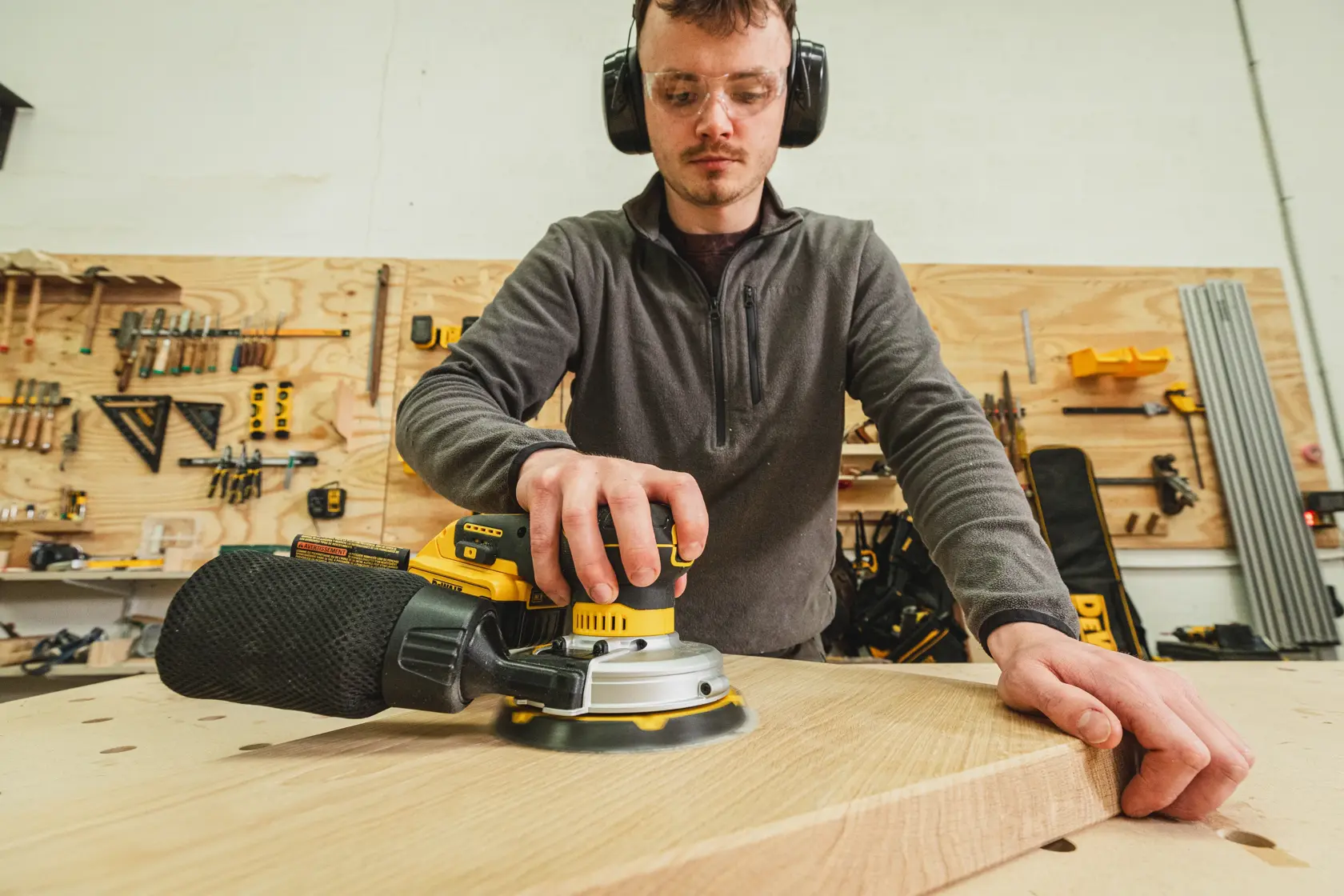 Personne utilisant une ponceuse DEWALT sur une planche en bois dans un atelier avec des outils en arrière-plan.