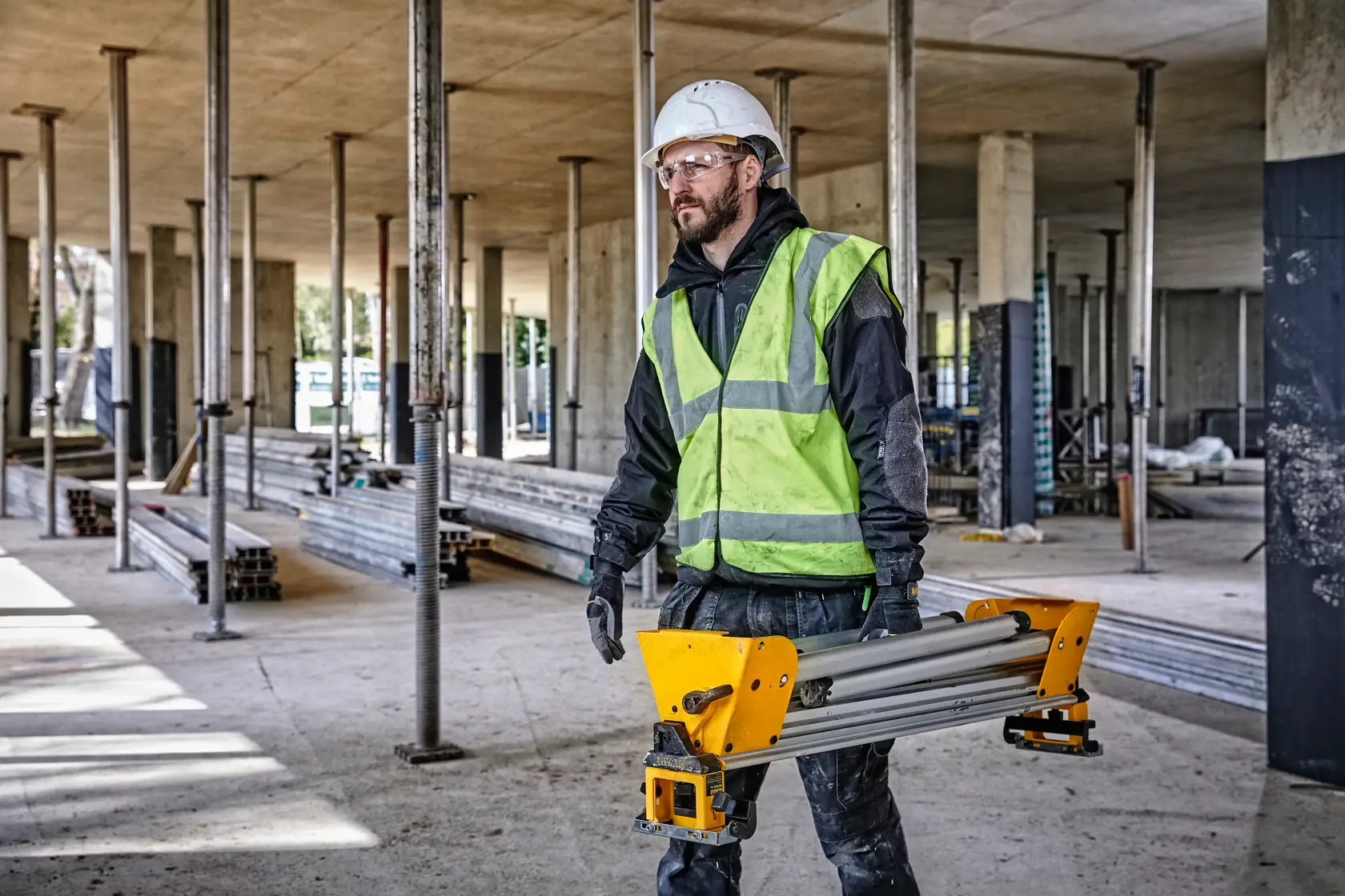 Ouvrier en gilet et casque porte un outil DEWALT DE7033 sur un chantier avec des poutres en acier.