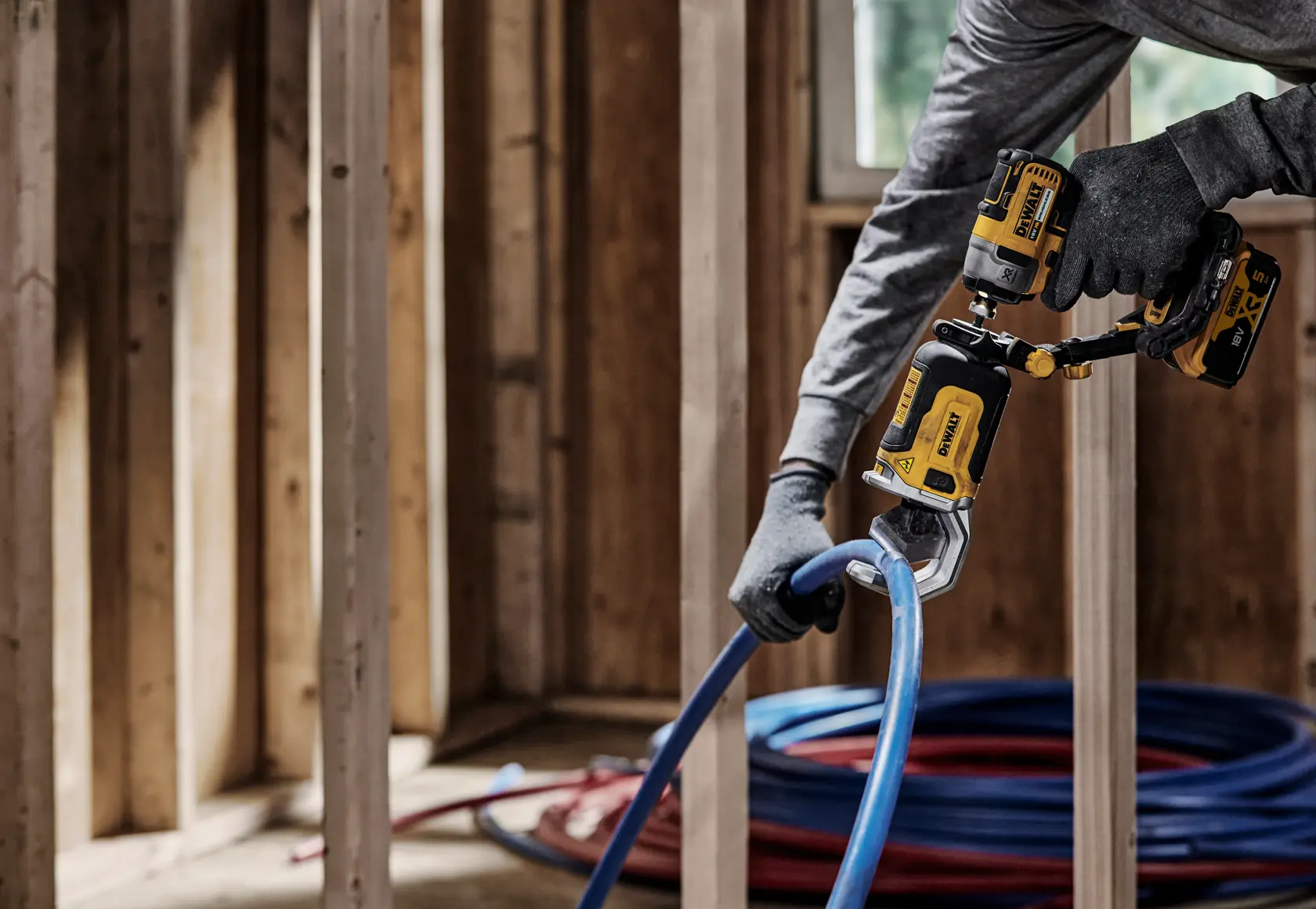 Una persona con guantes utiliza una herramienta eléctrica DEWALT DT20560 para cortar una tubería de plástico azul en una estructura interior de madera.