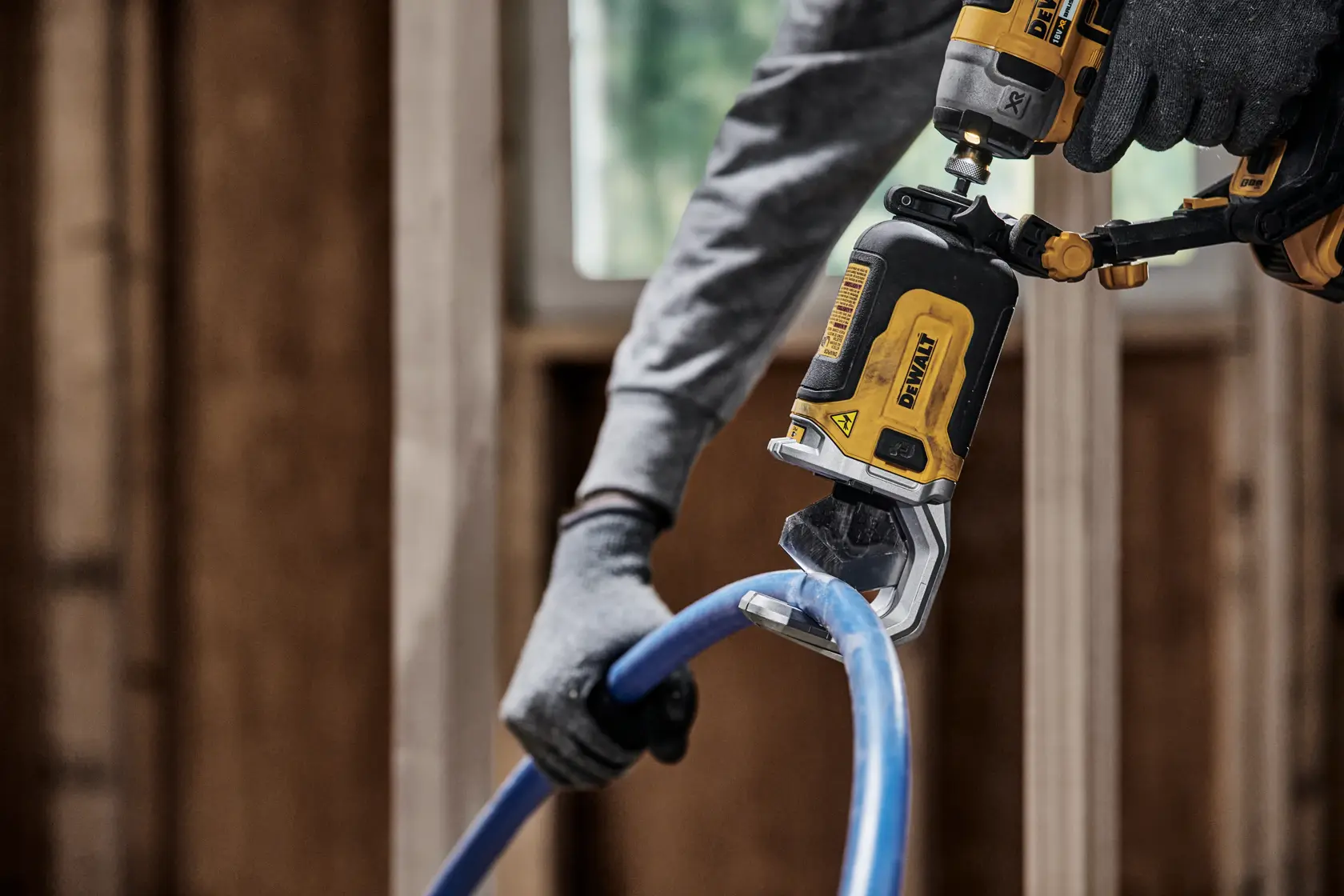 Una persona con guantes utiliza una herramienta eléctrica DEWALT DT20560 para cortar una tubería de plástico azul en un entorno de construcción.