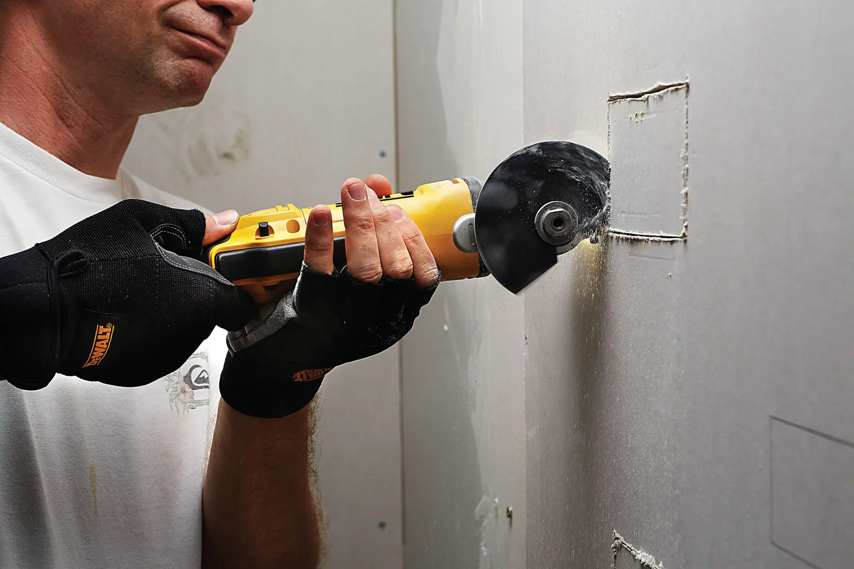 Une personne découpe un carré dans du placo avec un outil DEWALT et porte des gants noirs de protection.