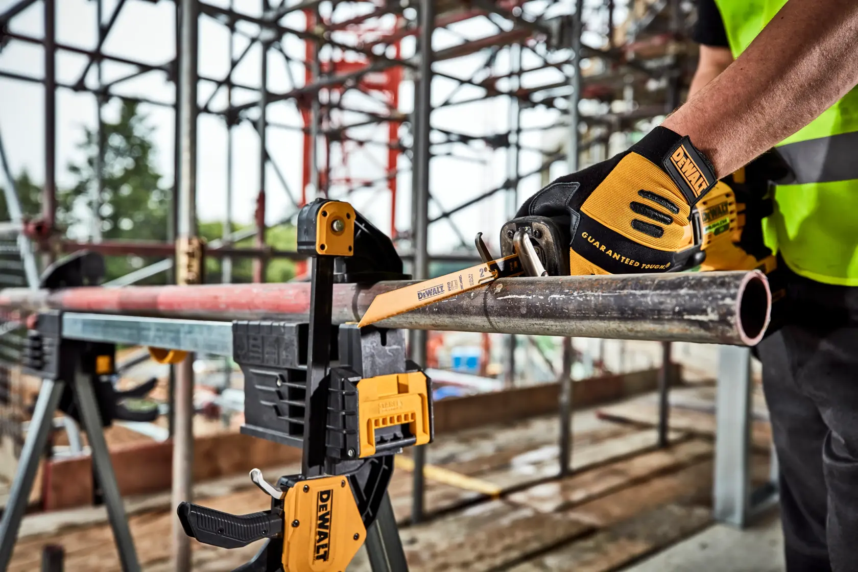 Une personne portant des gants DEWALT utilise une scie à main DEWALT pour couper un tube métallique, maintenu en place par un serre-joint DEWALT sur un chantier avec des échafaudages en arrière-plan.