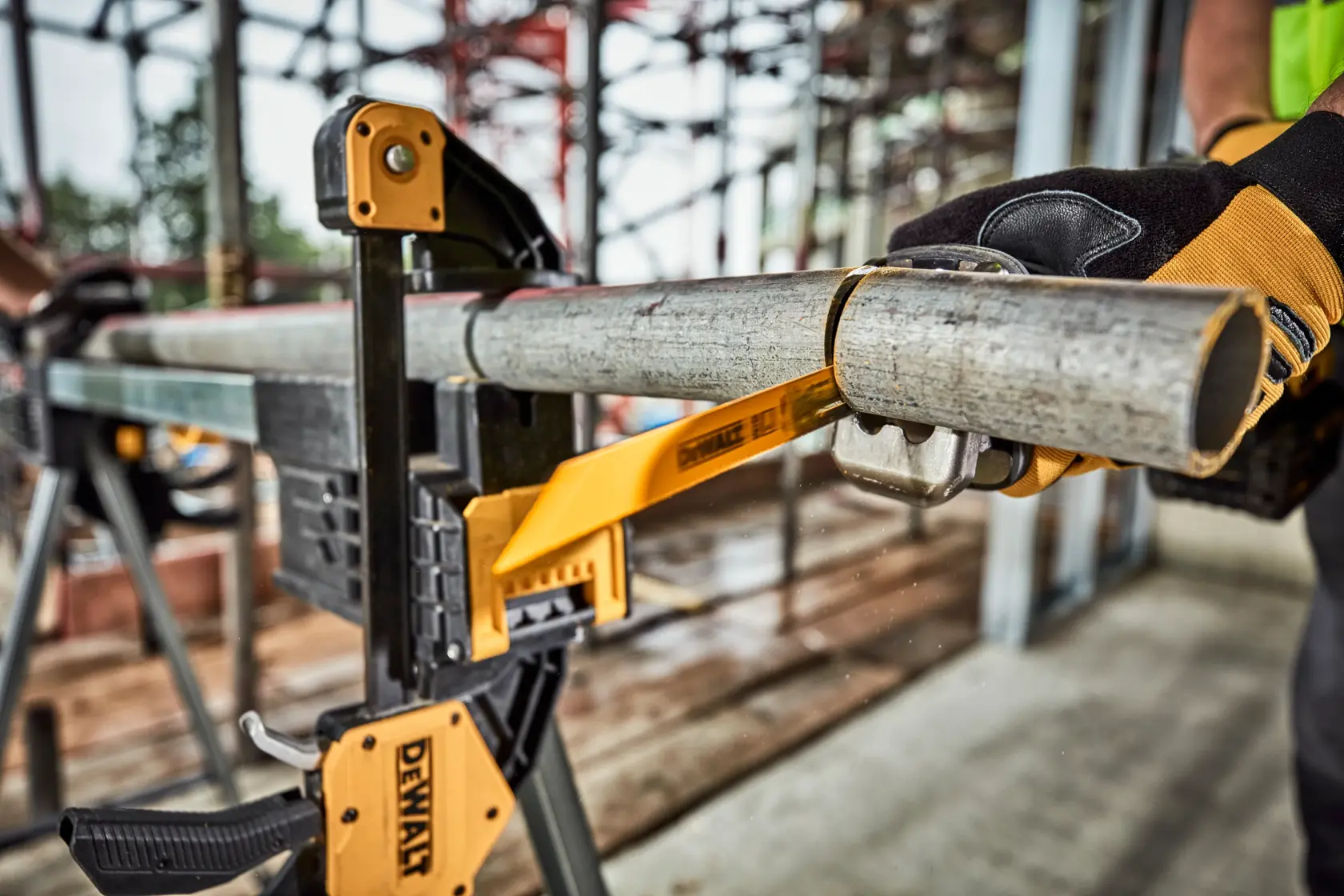 Une pince DEWALT maintient fermement un tuyau en métal sur un établi tandis qu'une personne portant des gants de protection utilise un coupe-tube pour couper le tuyau. L'environnement semble être un chantier de construction.